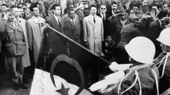 The image depicts a historical scene with a large crowd gathered, likely for a ceremonial or significant event. In the foreground, two soldiers are saluting while holding a flag, which features a crescent and a star, elements often associated with some national flags. The crowd appears to consist of men in various types of clothing, with some wearing military uniforms and others in suits. The overall atmosphere suggests a moment of national importance, possibly related to independence or a political event.