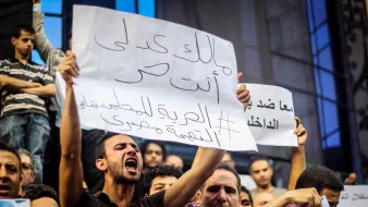 The image depicts a crowd of protestors holding signs during a demonstration. One individual is prominently featured, passionately expressing their views. The sign reads a message in Arabic, which translates to a call for freedom and justice. The crowd appears engaged and determined, highlighting the intense atmosphere of the protest. Various faces in the background show a mix of emotions, reflecting the seriousness of the situation.
