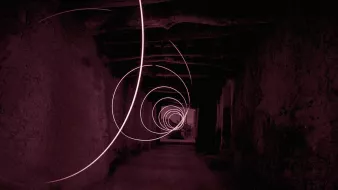The image depicts a long, seemingly dark corridor with a series of swirling light trails creating a spiral effect. The color tone is predominantly red, contributing to a mysterious and somewhat surreal atmosphere. The walls of the corridor appear rough and textured, enhancing the sense of depth and perspective as the light trails guide the eye towards the far end of the hallway. The overall composition evokes a feeling of motion and intrigue.