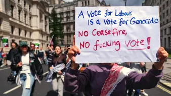 The image shows a protest scene with demonstrators in the background. In the foreground, a person is holding a sign that reads, "A Vote for Labour is a Vote for Genocide. NO Ceasefire, NO fucking VOTE!" The atmosphere appears to be charged with political activism, indicating strong sentiments regarding a current political issue.