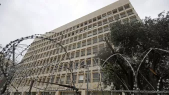 The image shows a large, modern building with a distinct architectural style, featuring multiple windows arranged in a grid pattern. In the foreground, there is a fence topped with barbed wire, indicating a level of security. The scene appears cloudy or overcast, giving a somewhat somber atmosphere. Additionally, there are some trees visible, possibly to the side of the building, adding a touch of greenery to the environment.