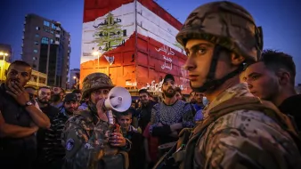 The image depicts a scene of a gathering in an urban environment during the evening. In the foreground, soldiers in military gear, including helmets, are present, with one holding a megaphone, suggesting a communication or announcement to the crowd. The background features a large, colorful wall or mural that displays the Lebanese flag and some Arabic text, likely conveying a political or social message. The crowd surrounding the soldiers appears engaged, with various expressions on their faces, indicating a sense of tension or anticipation in the atmosphere. The setting is illuminated by artificial lighting, hinting at the late hour.