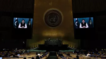 The image depicts a scene from a United Nations assembly. In the foreground, there are delegates seated at rows of desks, facing the main podium. The backdrop features a large screen displaying a speaker, who appears to be addressing the assembly. The setting is characterized by the assembly hall's distinctive architecture, featuring high walls and the emblem of the United Nations. The atmosphere is formal and legislative, reflecting an important discussion or speech taking place.