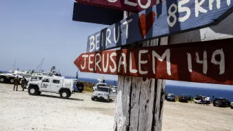 The image depicts a wooden signpost with several directional arrows pointing towards different locations. The names of the places, including "Beirut" and "Jerusalem," are painted in bold, colorful letters. The distances to these locations are also indicated in kilometers. In the background, there are vehicles, including a UN truck, parked on a sandy area near the coastline, with a view of the sea. The overall setting appears to be a scenic lookout or border area.