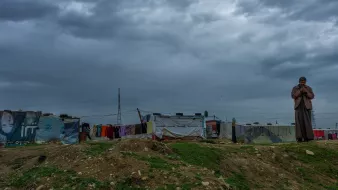 The image depicts a somber landscape under a cloudy sky. In the foreground, a lone figure stands, appearing pensive or contemplative. The background features makeshift structures, likely tents or shanties, indicating a temporary or informal settlement. These structures are adorned with various colors and materials, reflecting a harsh living environment. The overall atmosphere conveys a sense of solitude and resilience against the backdrop of an overcast sky.