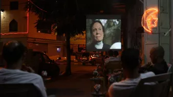 The image depicts a night scene where a group of people is seated in chairs, watching a film projected onto a large screen set up outside. The atmosphere appears relaxed, with some of the viewers enjoying drinks, possibly shisha. The area is illuminated by both the projection light and neon signage in the background, adding a lively ambiance to the setting. The scene captures a communal experience centered around outdoor movie viewing.