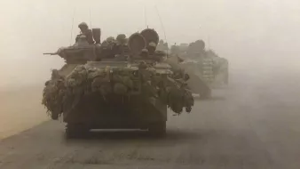 The image depicts military vehicles, specifically tanks, moving through a dusty and foggy environment. The tanks are heavily laden with gear and equipment, indicating they are prepared for deployment or combat. The visibility in the scene is limited due to the dust, which creates a muted atmosphere. Soldiers can be seen perched on top of the tanks, suggesting readiness and vigilance as they navigate the terrain. The overall mood conveys a sense of military operation amid challenging conditions.