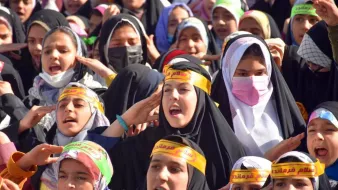 The image shows a large group of girls and young women gathered together, likely participating in an event or demonstration. They are wearing colorful headscarves and black clothing, along with yellow headbands that feature Arabic text. The girls appear to be actively engaged, singing or chanting, and expressing enthusiasm. Their expressions convey a sense of unity and purpose as they participate in this collective activity.