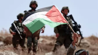 The image shows a man holding a Palestinian flag aloft, standing in a rocky landscape. He appears to be expressing a strong patriotic sentiment. In the background, a group of soldiers is visible, dressed in military gear, suggesting a tense or conflict-related scene. The setting looks arid, typically associated with certain regions in Palestine. The overall atmosphere conveys a sense of tension and resistance.