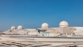 The image shows a large industrial facility, likely a nuclear power plant, with several prominent dome-shaped structures. The plant features a modern design with various buildings and infrastructure surrounding it, set against a clear blue sky. The setting appears to be an arid or semi-arid environment, with minimal vegetation. There are parking areas and some infrastructure elements visible in the foreground, indicating it is a well-developed site.