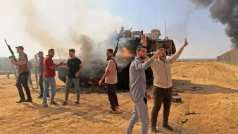 The image depicts a chaotic scene in a sandy area where a group of individuals appears to be celebrating or protesting near a burning military vehicle. Smoke rises from the vehicle, creating a dramatic backdrop. Some people are visible holding firearms, while others are raising their hands, possibly in a gesture of victory. The environment suggests a conflict or demonstration, with a tense atmosphere reinforced by the presence of smoke and the military vehicle. The landscape is open, with a barrier visible in the background, indicating a border or contested area.