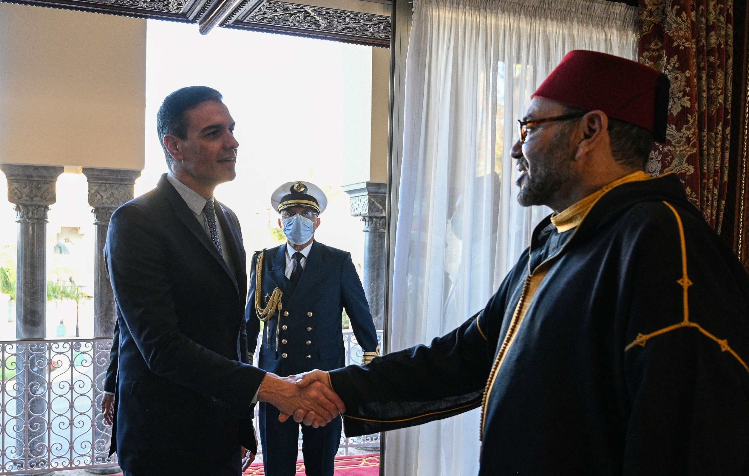 La imagen muestra a dos hombres dándose la mano en un entorno formal. Uno parece ser un líder o dignatario, vestido con un traje oscuro, mientras que el otro lleva una vestimenta tradicional, posiblemente un manto y un fez. En el fondo, se puede ver a un tercero, que es un guardia o oficial en uniforme, observando la escena. La decoración del lugar es elegante, con cortinas blancas y un entorno arquitectónico notable.