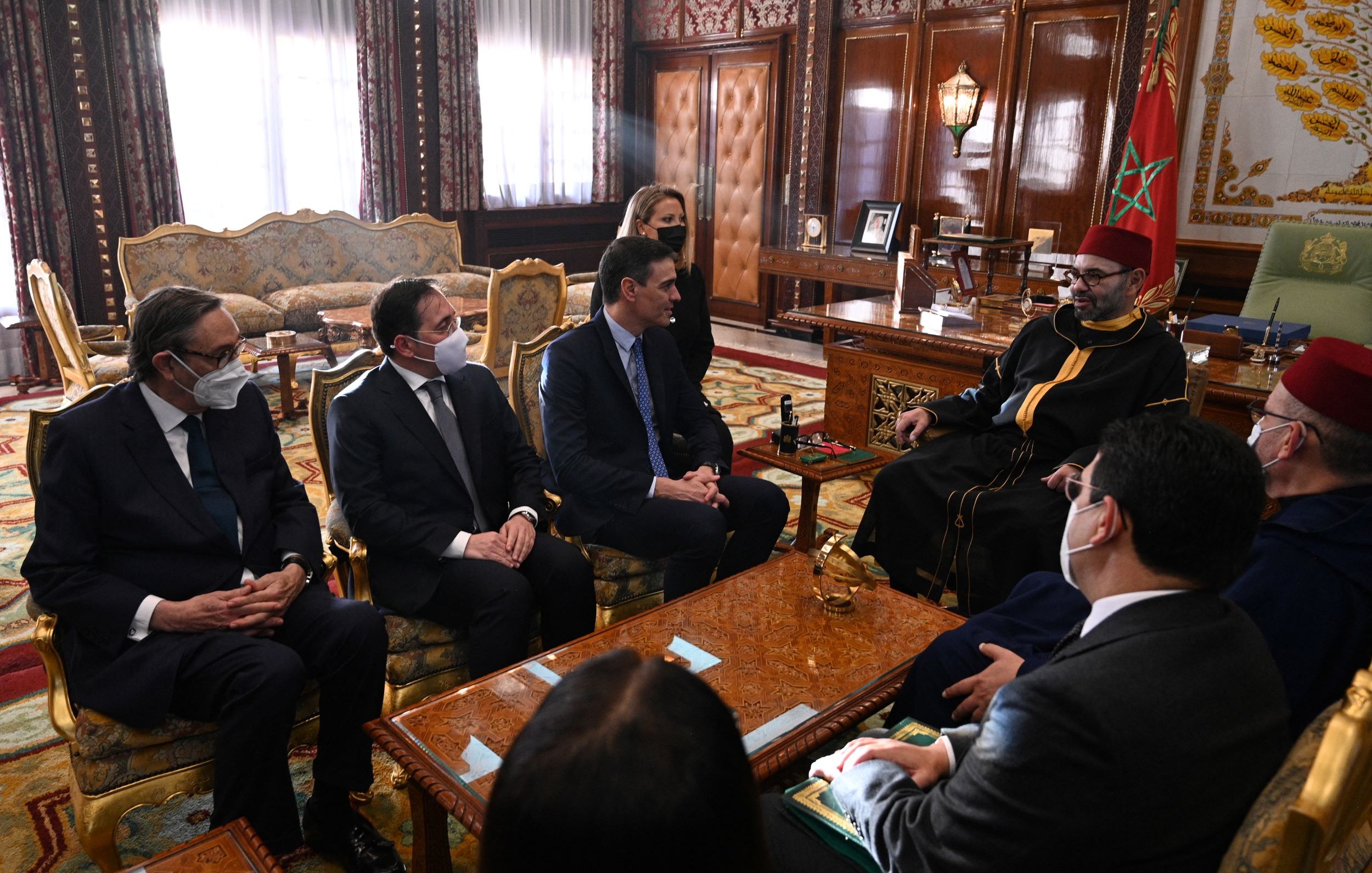 La imagen muestra una reunión en un ambiente formal y lujoso, posiblemente en un palacio. Hay varias personas sentadas alrededor de una mesa. Algunos de los asistentes llevan puesto un tapabocas, lo que sugiere que la reunión puede haber tenido lugar durante un periodo de preocupación por la salud pública, como la pandemia. En el centro de la escena, hay un individuo que parece ser una figura de autoridad, vestido con túnica y gorro tradicional. El interior está decorado con un estilo opulento, que incluye una alfombra elaborada y muebles de alta calidad. Las expresiones de los asistentes son serias, indicando la importancia del encuentro.