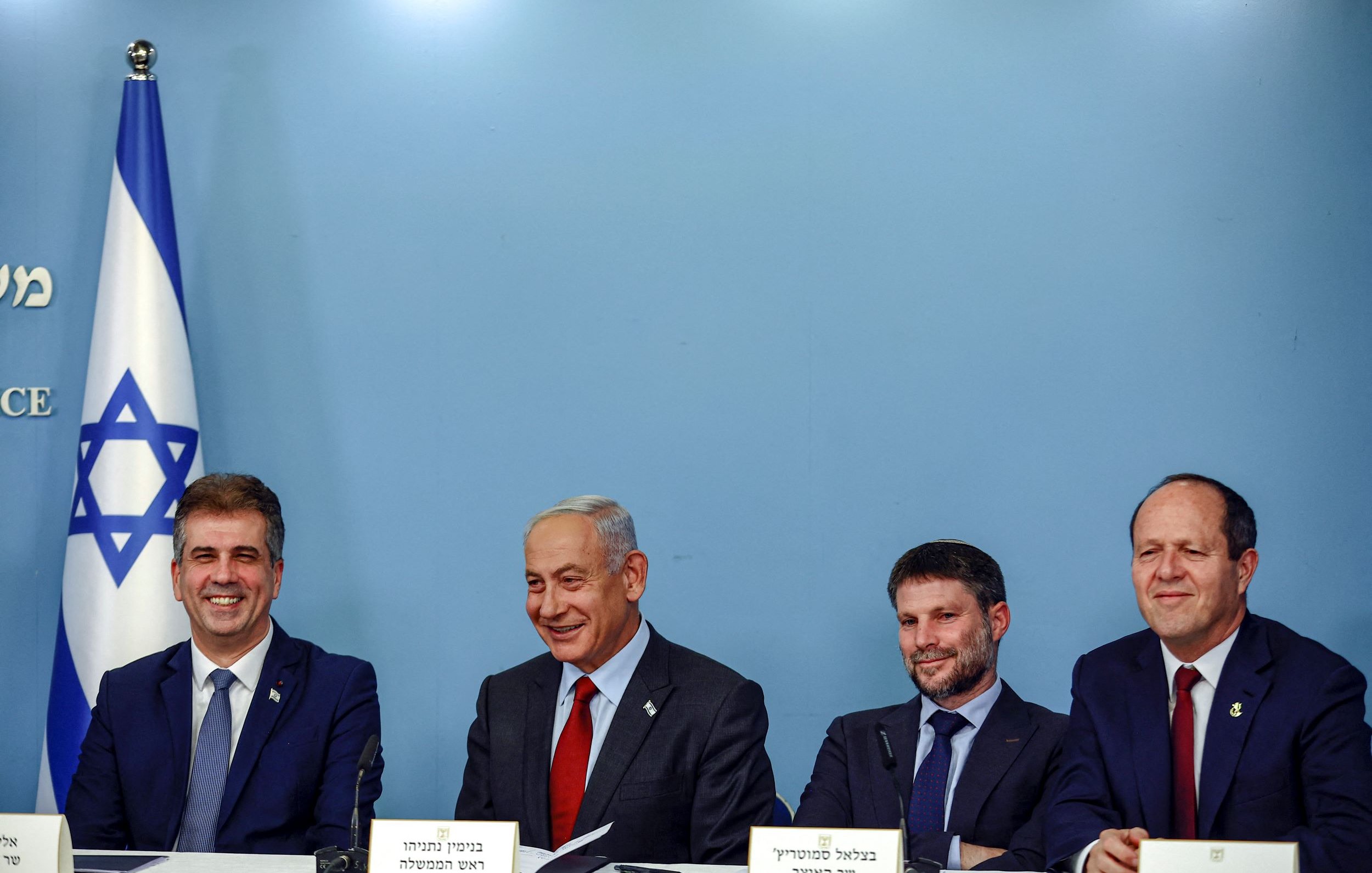 La imagen muestra a un grupo de cuatro personas sentadas alrededor de una mesa en un entorno oficial. Al fondo, se puede ver una bandera de Israel. Las personas parecen estar en un evento o una conferencia, y todos tienen expresiones serias pero amigables. Están vestidos de manera formal. La escena sugiere un contexto político o gubernamental.