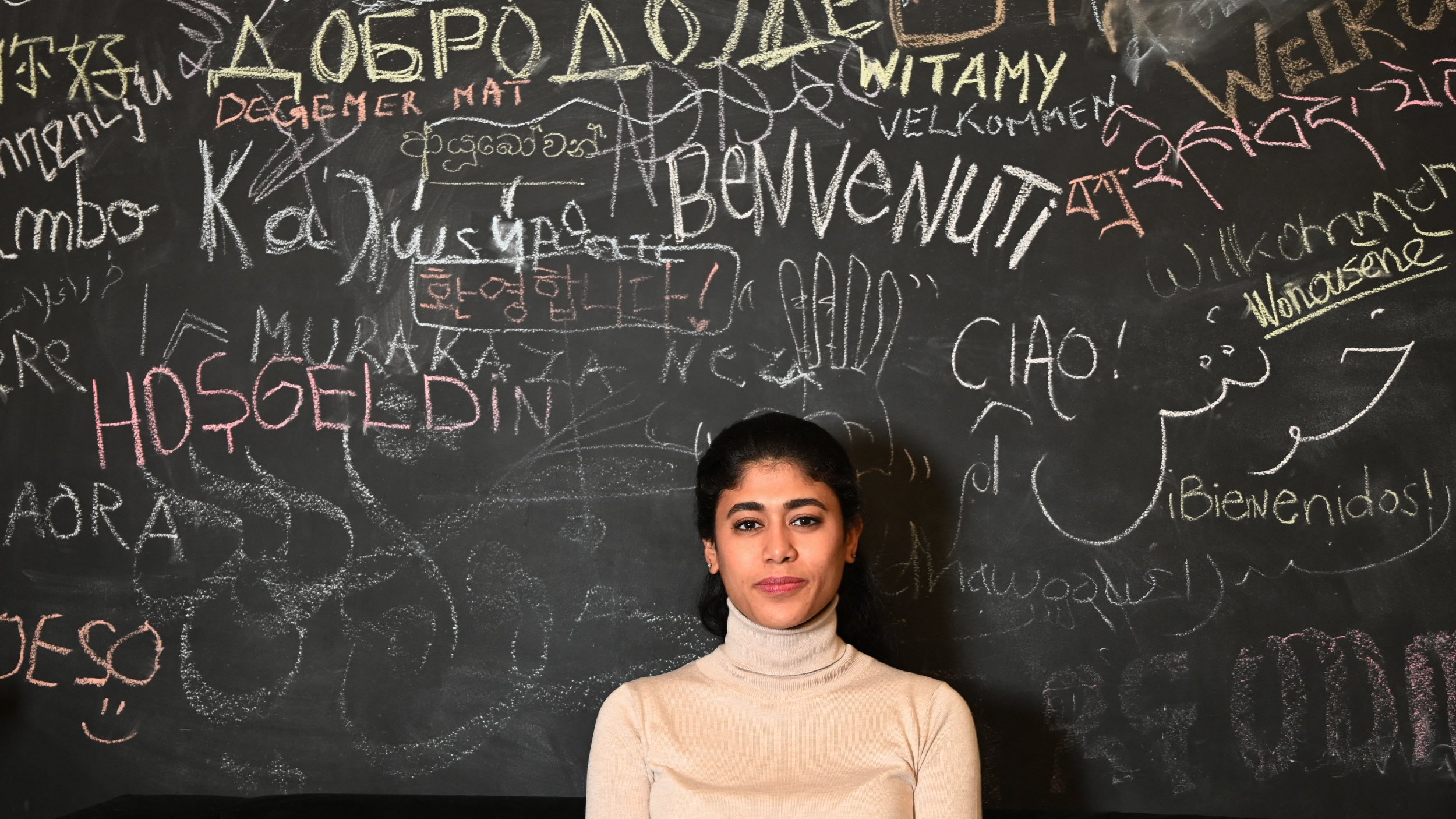 La imagen muestra a una joven posando frente a una pared de tiza oscura cubierta de múltiples palabras y frases escritas en diferentes idiomas. La mujer lleva un suéter claro y tiene el cabello recogido. La pared refleja un ambiente multicultural, con saludos y palabras de bienvenida en distintas lenguas.