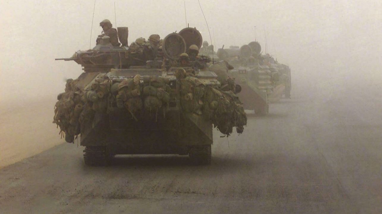 La imagen muestra un convoy militar en un ambiente polvoriento y desértico. Se pueden ver varios vehículos blindados, posiblemente tanques, avanzando en formación. Los vehículos están cargados con equipos y suministros, lo que sugiere que están en una misión de combate o de suministro. La visibilidad es limitada debido a la niebla o polvo, lo que añade una atmósfera tensa a la escena.