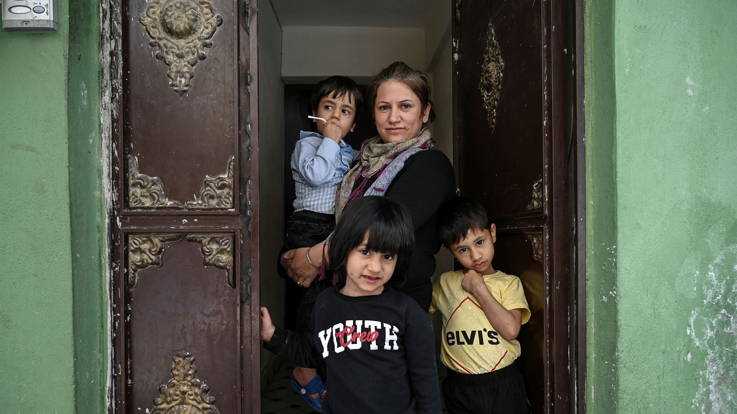 En la imagen se puede ver a una mujer de pie en una puerta, sosteniendo un cigarrillo y rodeada por tres niños. Dos de los niños están detrás de ella, mientras que uno se encuentra al frente. La mujer parece estar sonriendo levemente, y el ambiente parece ser de un lugar residencial, con paredes verdes y una puerta antigua adornada. Los niños tienen expresiones curiosas y están vestidos con ropa casual. La escena transmite una atmósfera familiar y cercana.