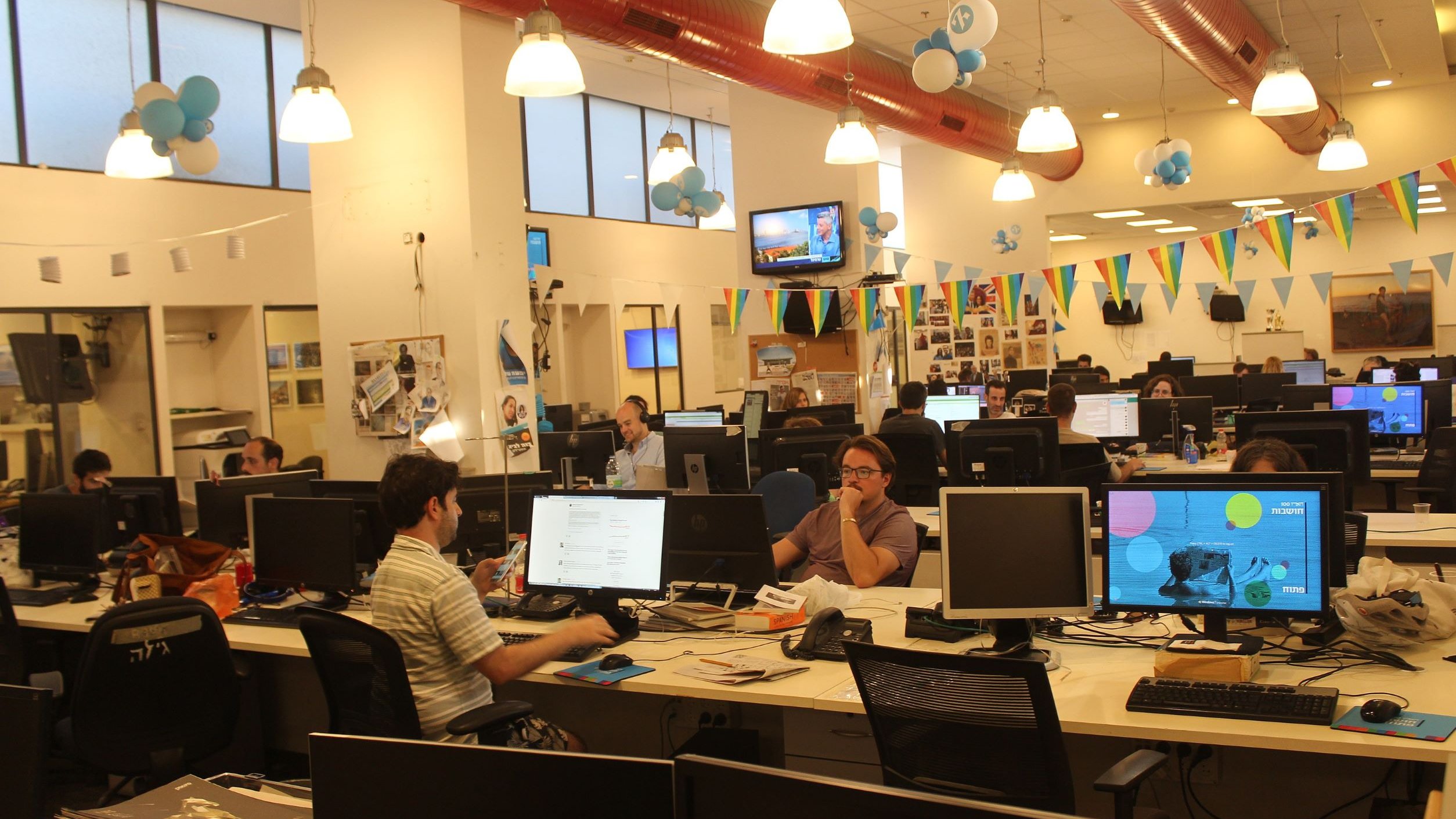 La imagen muestra una oficina moderna y espaciosa. Hay varias estaciones de trabajo con computadoras y pantallas en escritorios. Algunas personas están concentradas en sus tareas mientras que otras parecen colaborar o comunicarse. El ambiente es iluminado y decorado con globos y banderas, lo que sugiere que podría ser un lugar festivo o creativo. Las paredes tienen fotografías y otros elementos decorativos, añadiendo un toque personal al espacio laboral.