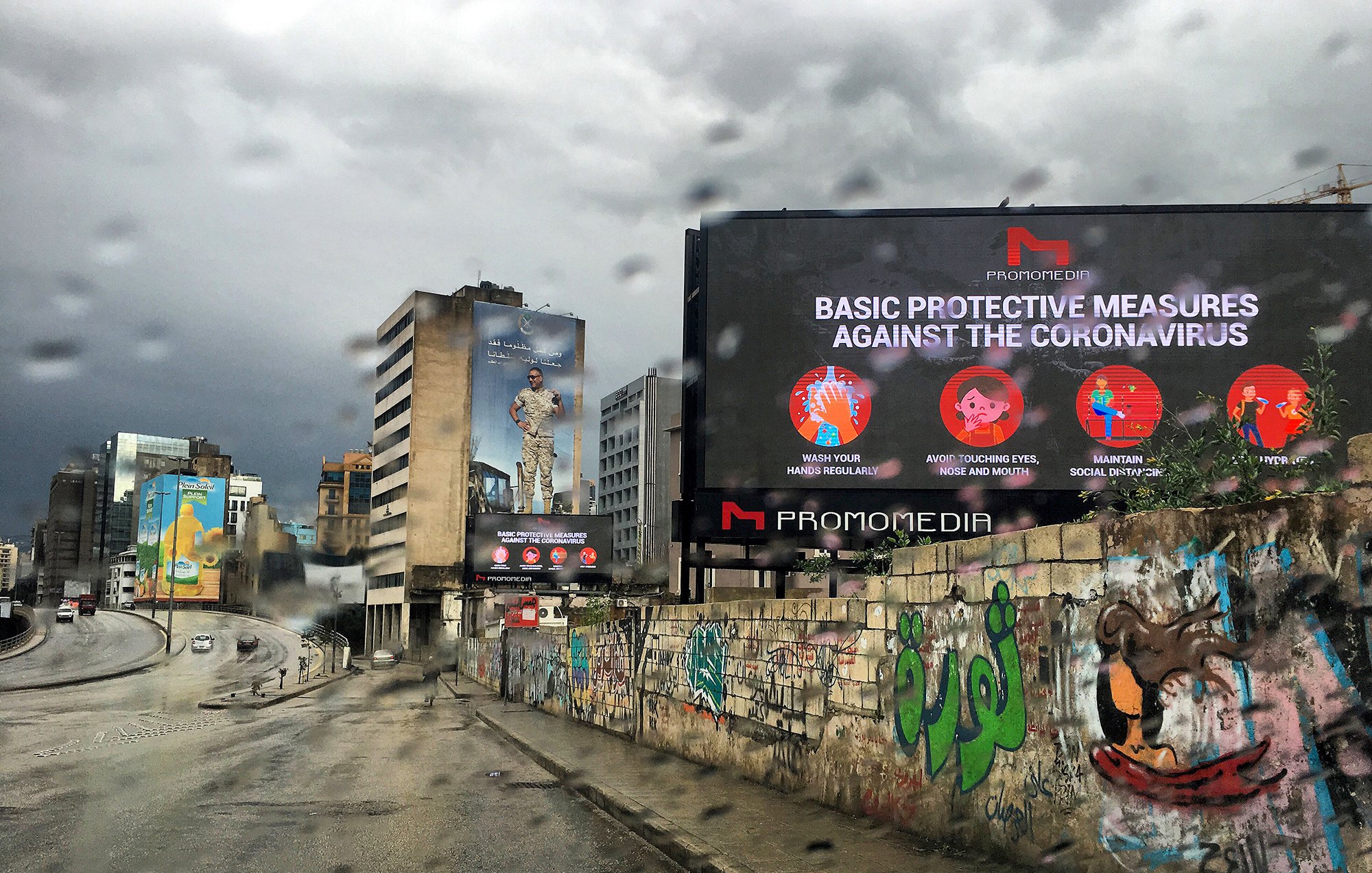 La imagen muestra una calle en un entorno urbano, con un ambiente lluvioso. Hay un letrero digital grande que presenta medidas de protección básicas contra el coronavirus. En el fondo, se pueden ver edificios y murales coloridos en las paredes, lo que añade un contraste visual a la escena. El cielo está nublado, lo que sugiere un clima gris y lluvioso.