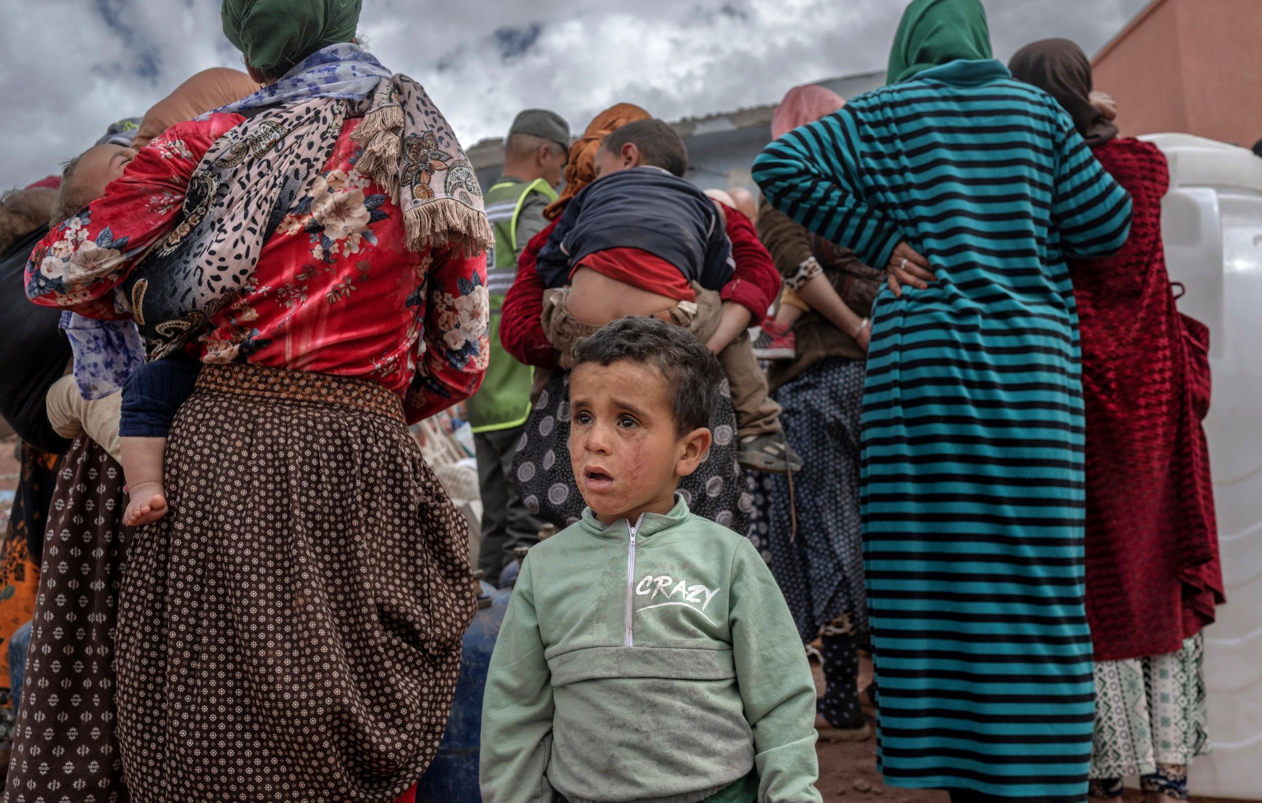 La imagen muestra a un grupo de personas de espaldas, predominantemente mujeres, que parecen estar en un ambiente rural. La ropa que llevan es variada, con patrones y colores diversos. En el primer plano, un niño pequeño aparece al frente, mirando hacia la cámara con una expresión que podría reflejar confusión o preocupación. El fondo sugiere un cielo nublado y la escena evoca una sensación de comunidad y un posible evento o reunión.