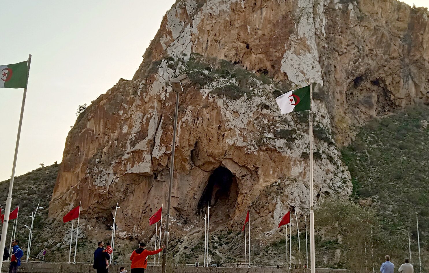 La imagen muestra un paisaje montañoso con una gran roca o acantilado en el centro, que tiene una cueva visible en su base. A su alrededor, hay varias banderas ondeando, entre las cuales algunas son de Argelia, ya que tienen un diseño distintivo. En la parte inferior de la imagen, se pueden ver algunas personas caminando y disfrutando del lugar. El cielo tiene un tono claro, posiblemente al atardecer. La escena refleja un ambiente natural y un sitio de interés cultural o histórico.
