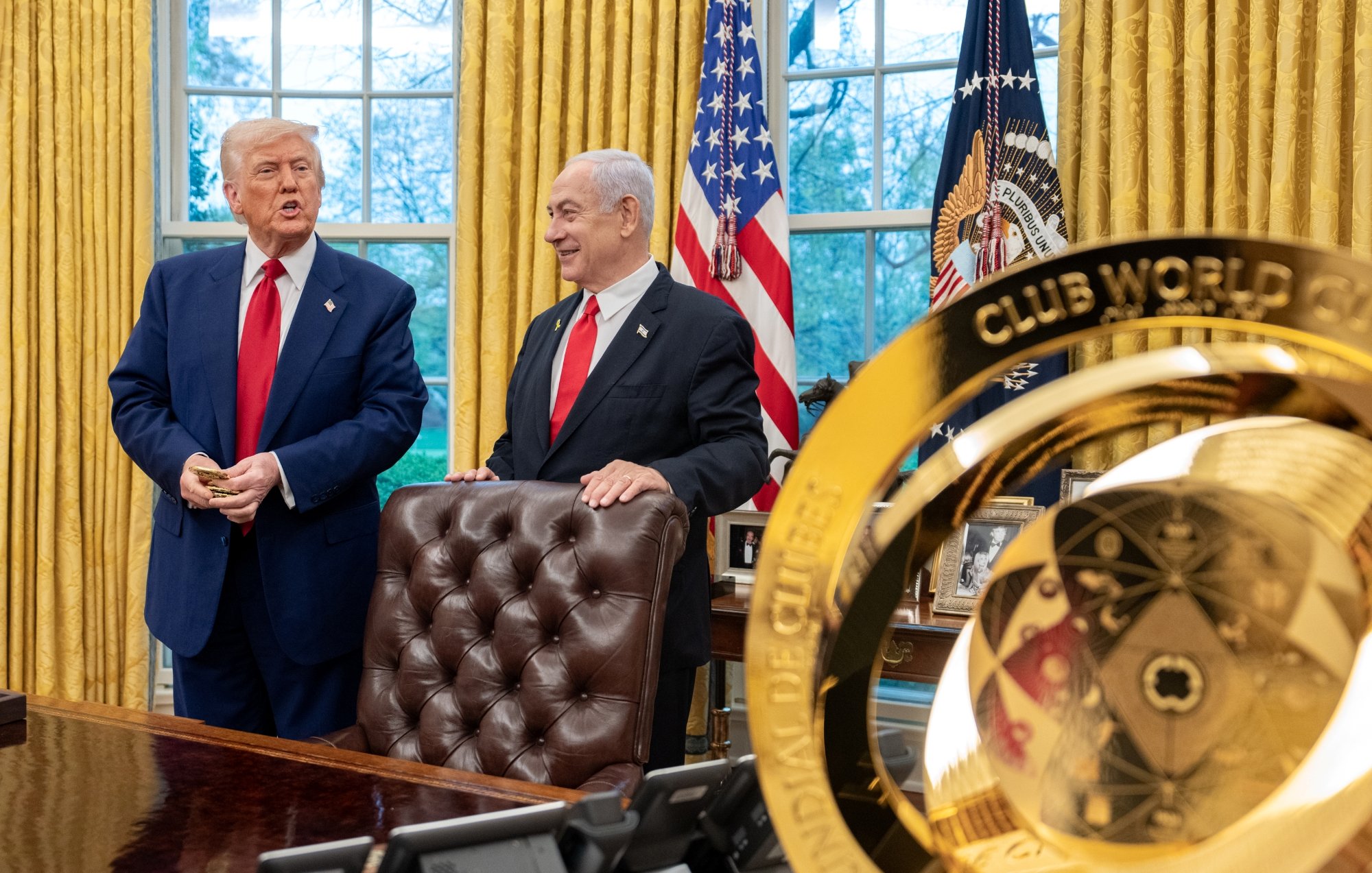 Two men in suits stand in an ornate office with gold curtains and flags, one holding a small object.