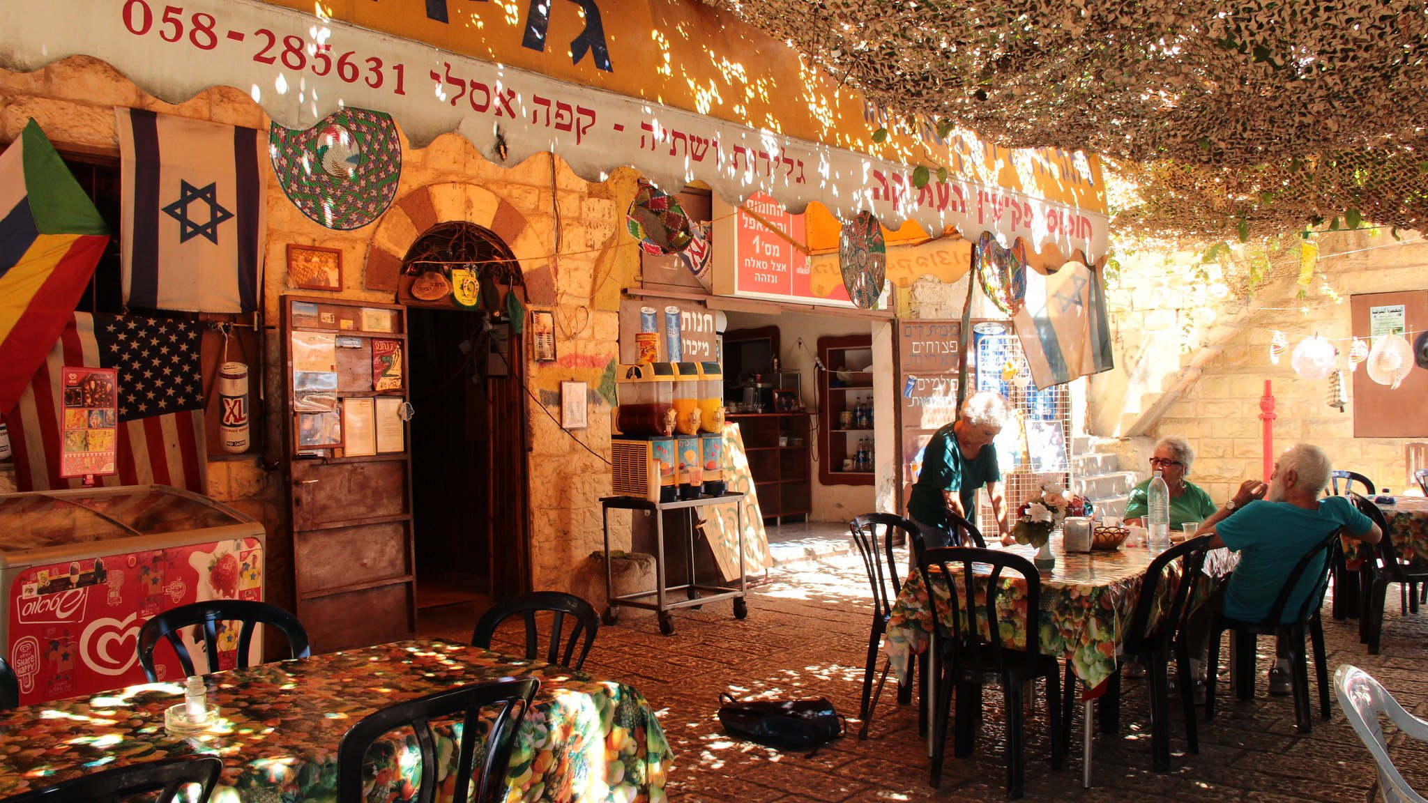 La imagen muestra un ambiente acogedor y colorido de un café o restaurante al aire libre. Hay mesas y sillas negras distribuidas sobre un suelo pavimentado, y la decoración incluye manteles con estampados florales. En el fondo, se pueden ver varias banderas, incluyendo la de Israel, colgadas en las paredes de piedra. El espacio está cubierto por una especie de toldo que filtra la luz, creando un ambiente agradable. Algunas personas están sentadas en las mesas, disfrutando del lugar.