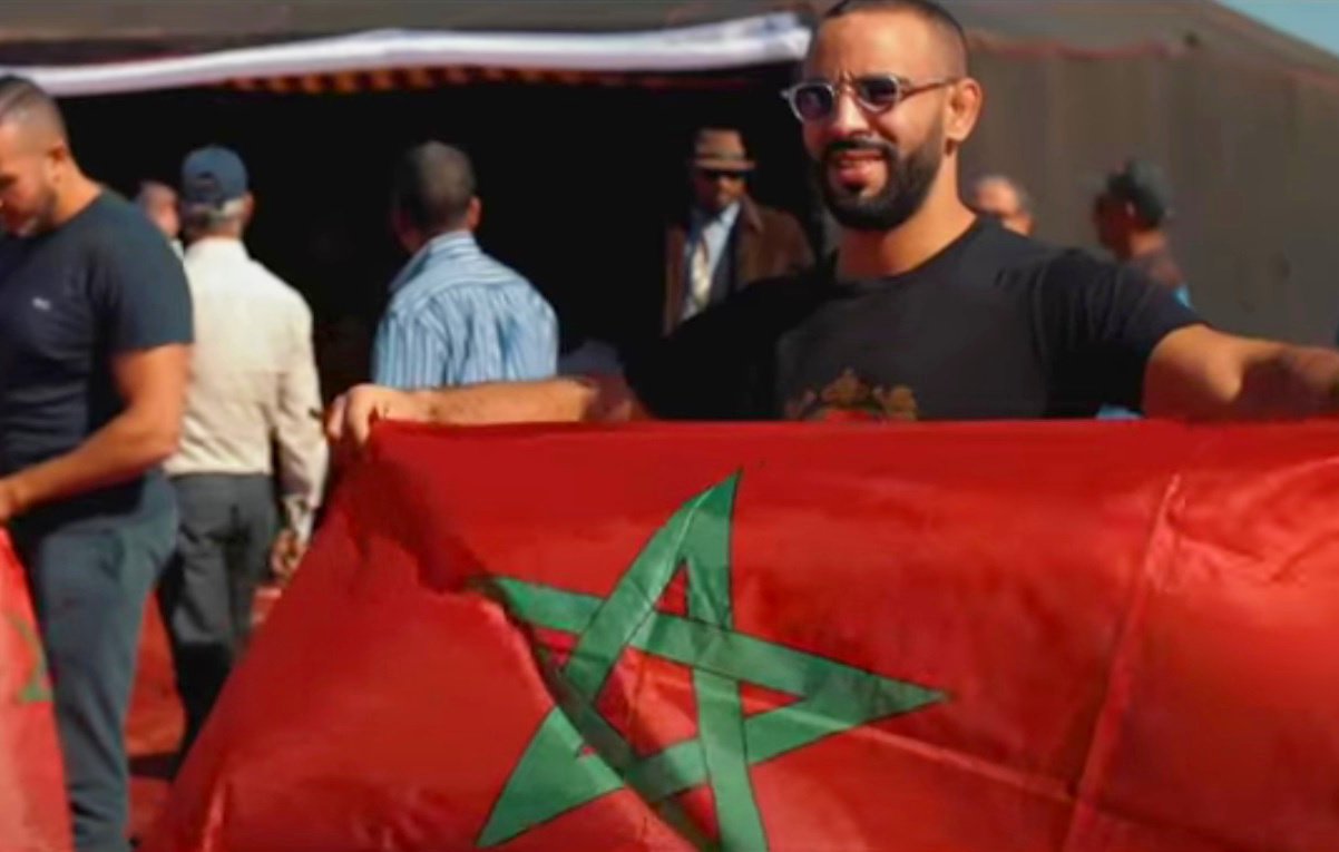 En la imagen se puede ver a un grupo de personas en un ambiente al aire libre, posiblemente en un evento o celebración. Un hombre sostiene una bandera de Marruecos, que es de color rojo con una estrella verde en el centro. Otros hombres también están presentes en el fondo, algunos sosteniendo banderas o participando en la actividad. La escena refleja un ambiente de camaradería y orgullo nacional.