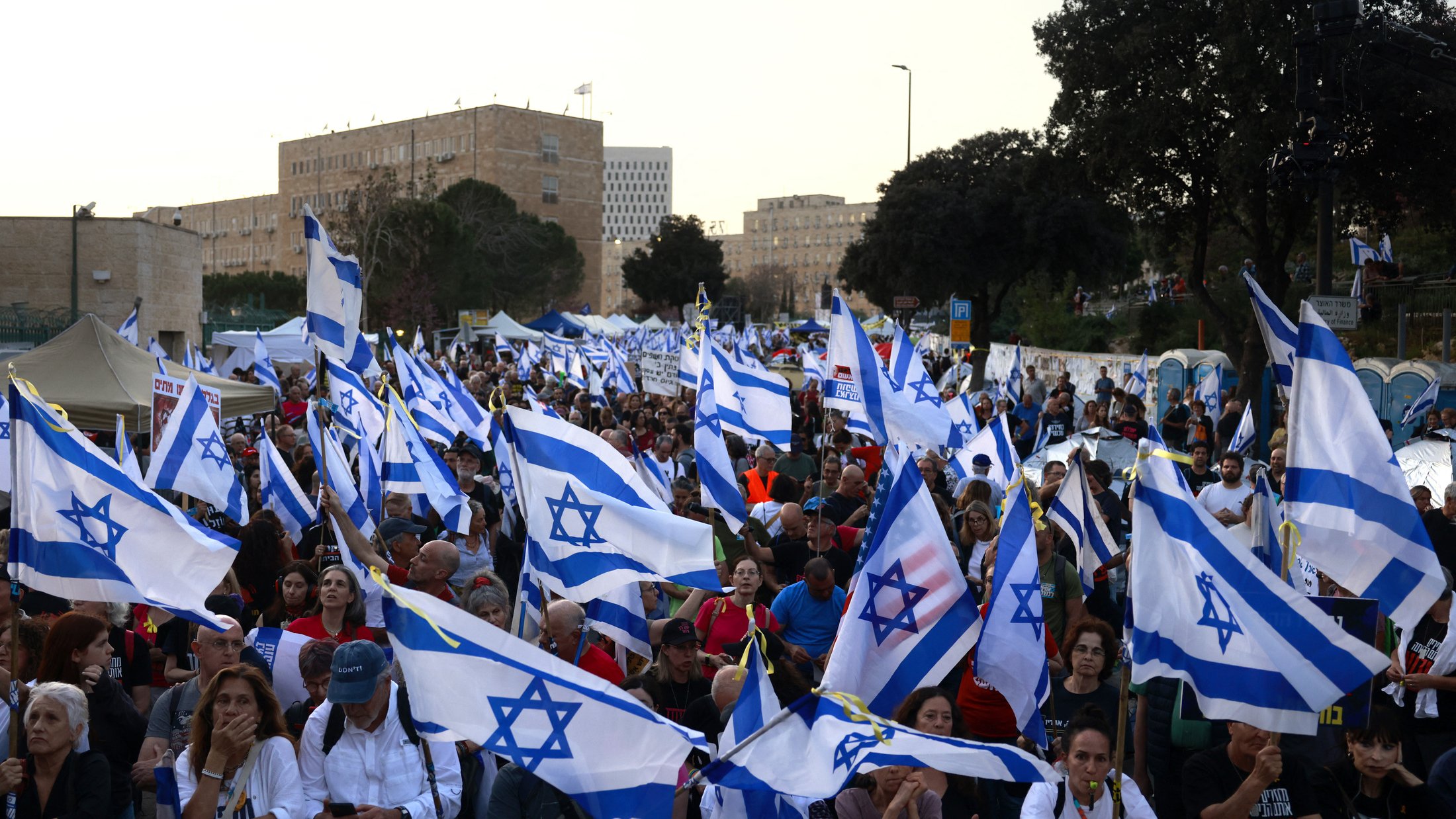 La imagen muestra una multitud de personas en una manifestación, ondeando banderas de Israel. La escena parece vibrante y llena de energía, con una gran cantidad de asistentes que levantan las banderas en un ambiente de protesta o celebración. En el fondo se pueden apreciar árboles y estructuras, lo que sugiere que el evento tiene lugar al aire libre, probablemente en una ciudad. La multitud parece diversa y está unida por una causa común.