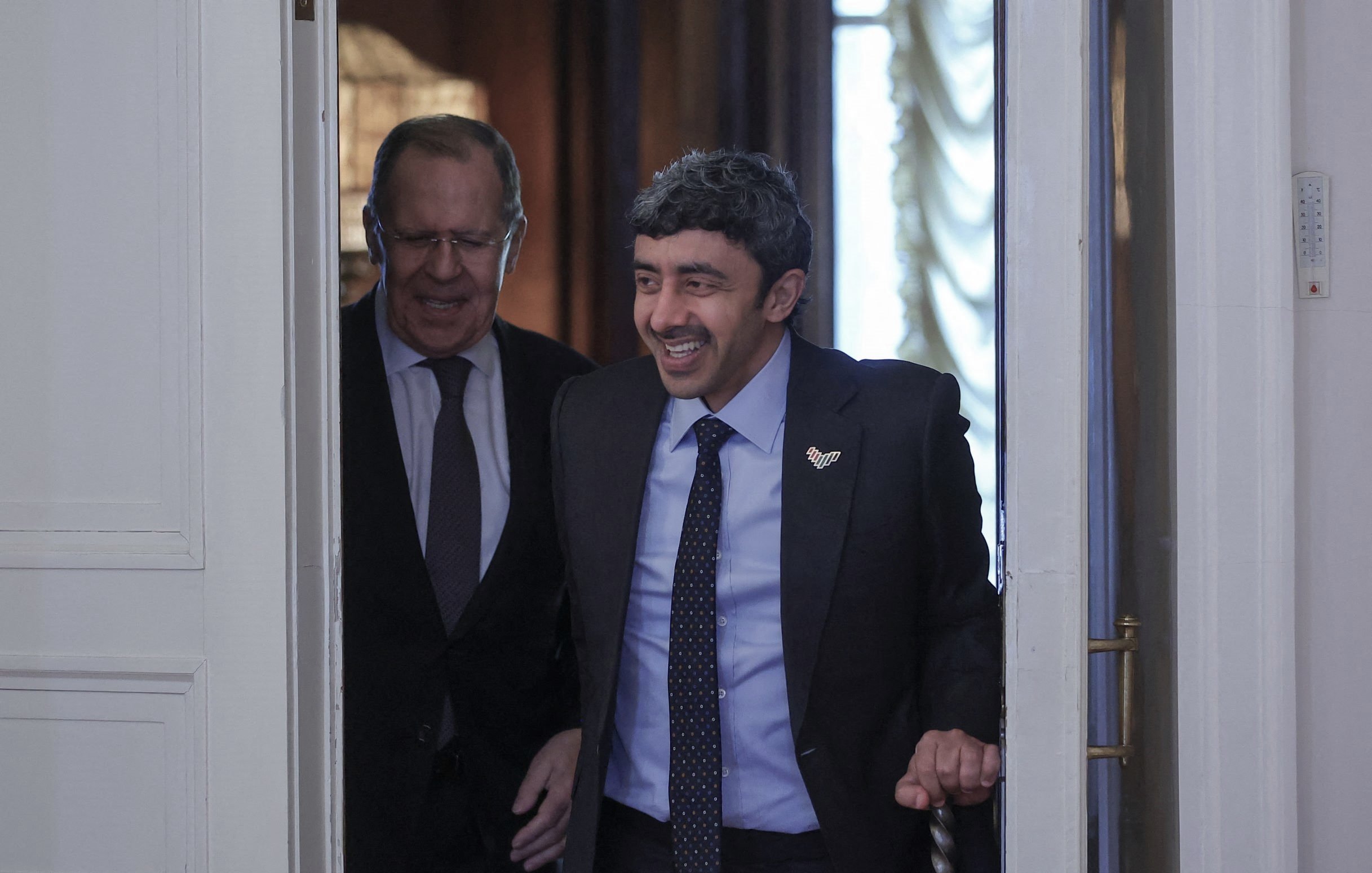 La imagen muestra a dos hombres entrando en una sala. Uno de ellos tiene una sonrisa amplia y está vestido con un traje oscuro y una corbata con un patrón. El otro hombre parece estar sonriendo también y lleva unas gafas. La decoración del fondo sugiere un ambiente formal y elegante, con luces suaves y detalles arquitectónicos. Ambos parecen estar en un momento de cordialidad y alegra.