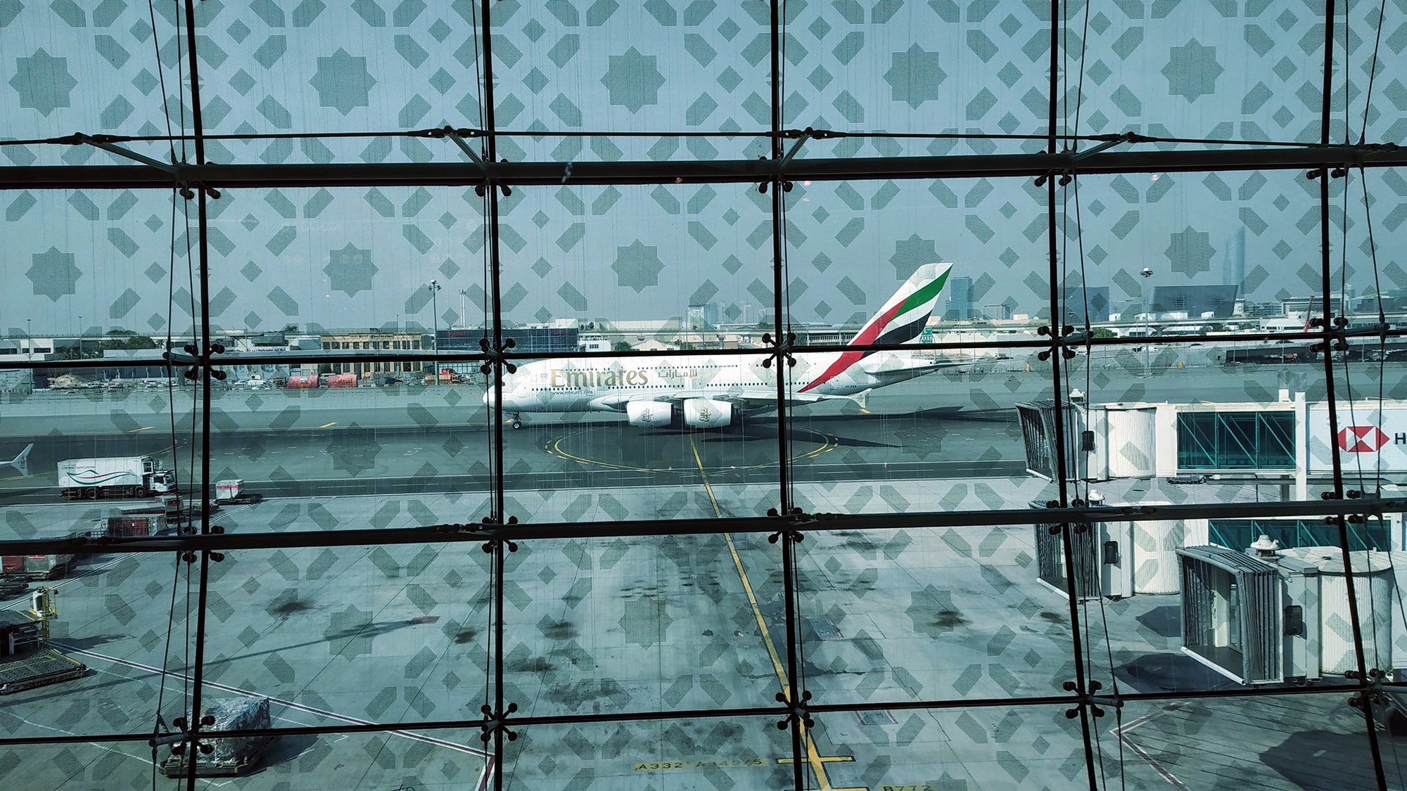 La imagen muestra la vista desde el interior de un aeropuerto, donde se puede ver un avión de la aerolínea Emirates estacionado en la pista. Hay una estructura de cristal en primer plano que tiene un patrón decorativo. En el fondo, se pueden observar otras áreas del aeropuerto y algunos vehículos de servicio. La atmósfera parece luminosa y abierta, típica de un gran terminal aéreo.