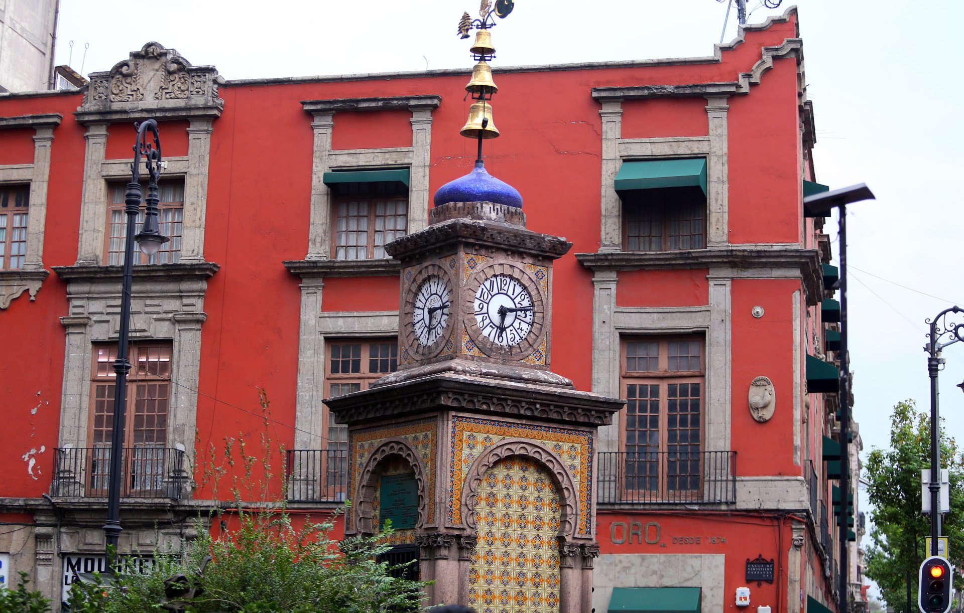 La imagen muestra un hermoso reloj de torre ubicado en una calle. La torre tiene una estructura ornamentada con un diseño colorido en su base y dos relojes visibles en los lados. Encima del reloj, hay una veleta dorada en la parte superior. Al fondo, se puede ver un edificio de color rojo con ventanas con contraventanas verdes. La arquitectura es de estilo colonial, con detalles decorativos en las fachadas. También hay una vegetación ligera en primer plano.