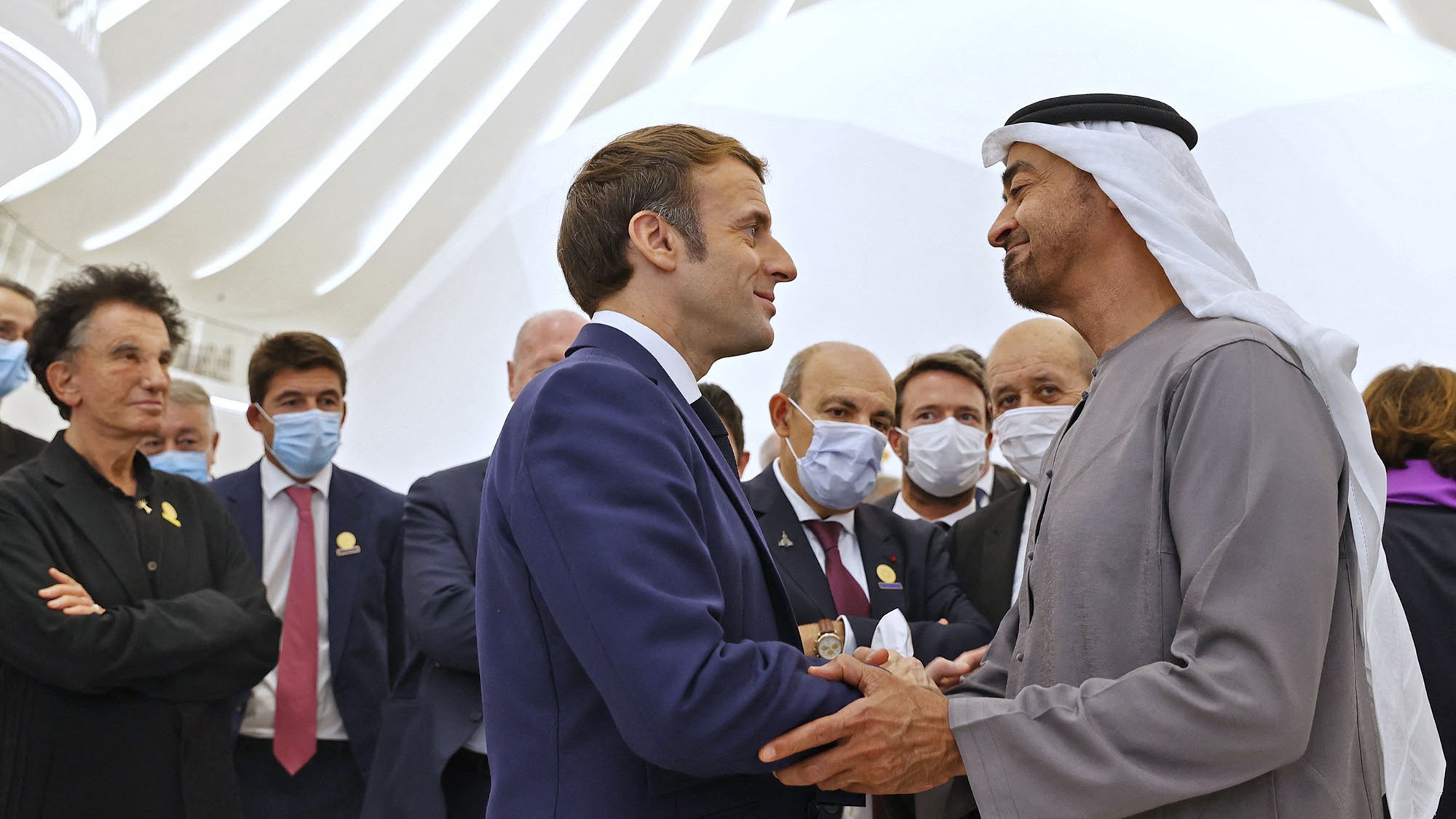 En la imagen se pueden ver a varias personas en un ambiente formal. Al frente, dos hombres están estrechando manos, uno de ellos viste un traje y el otro lleva una vestimenta tradicional árabe. Alrededor de ellos, hay más personas que observan la interacción, algunas de las cuales llevan mascarillas. El fondo muestra una arquitectura moderna y luminosa. La atmósfera es de cordialidad y diplomacia.