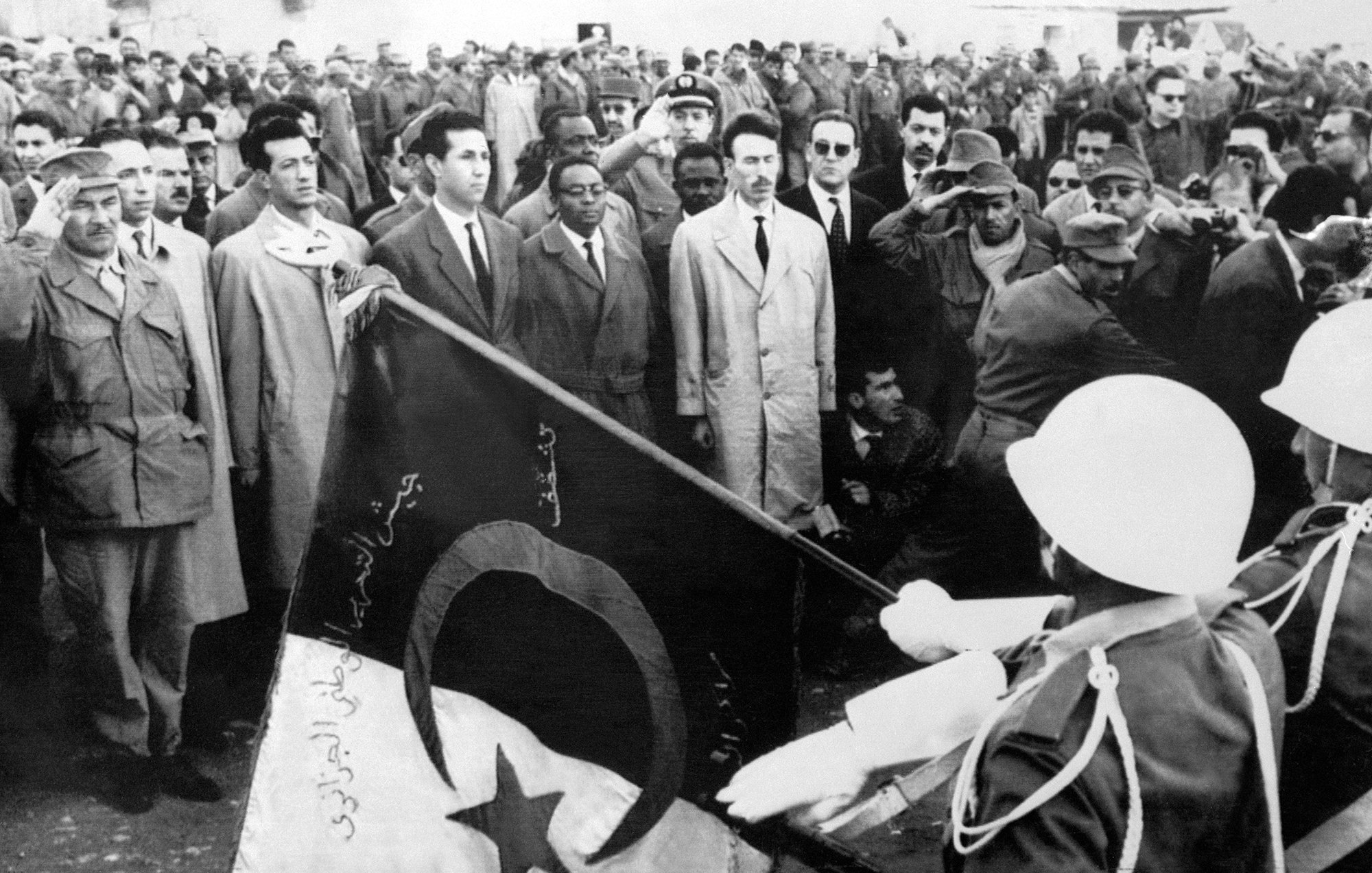 La imagen muestra un evento significativo, donde un grupo de personas se congrega. En el primer plano, soldados sostienen una bandera con símbolos distintivos, mientras que un grupo de hombres observa atentamente, algunos de ellos en posiciones de saludo. El ambiente parece ser solemne y formal, posiblemente relacionado con un momento histórico o ceremonioso. La vestimenta de los asistentes varía, con algunos en trajes y otros en uniformes militares, lo que sugiere una mezcla de civil y militar en el evento.