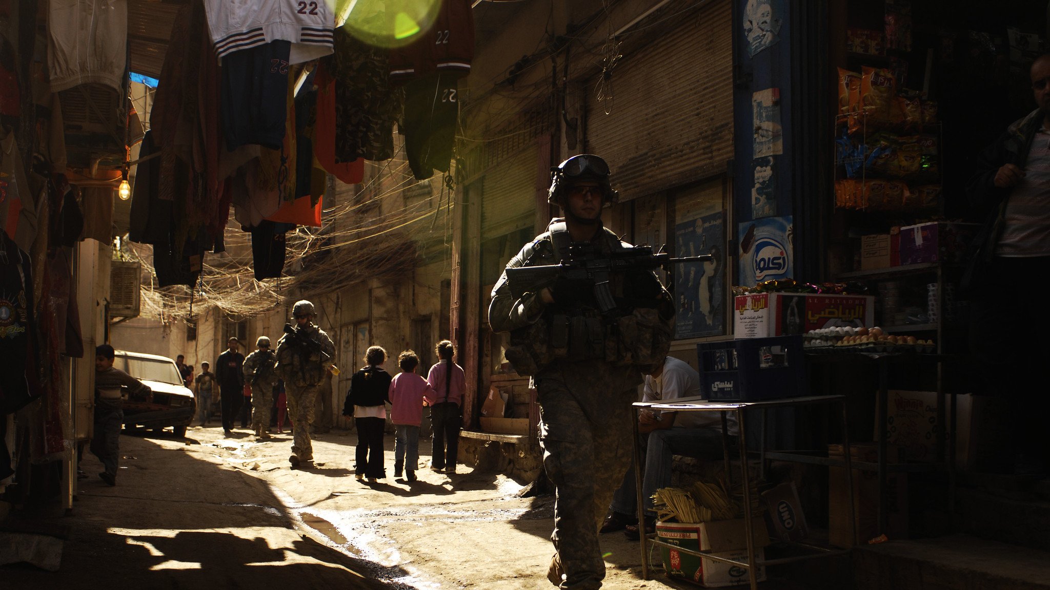 La imagen muestra a soldados patrullando en un callejón estrecho y iluminado por la luz del sol. A los lados, hay ropa colgada y tiendas de barrio que parecen ofrecer diversos productos. Un grupo de niños está presente en la escena, observando a los soldados. La atmósfera es tensa, dado el contexto militar, pero la luz crea un efecto visual interesante en el entorno.