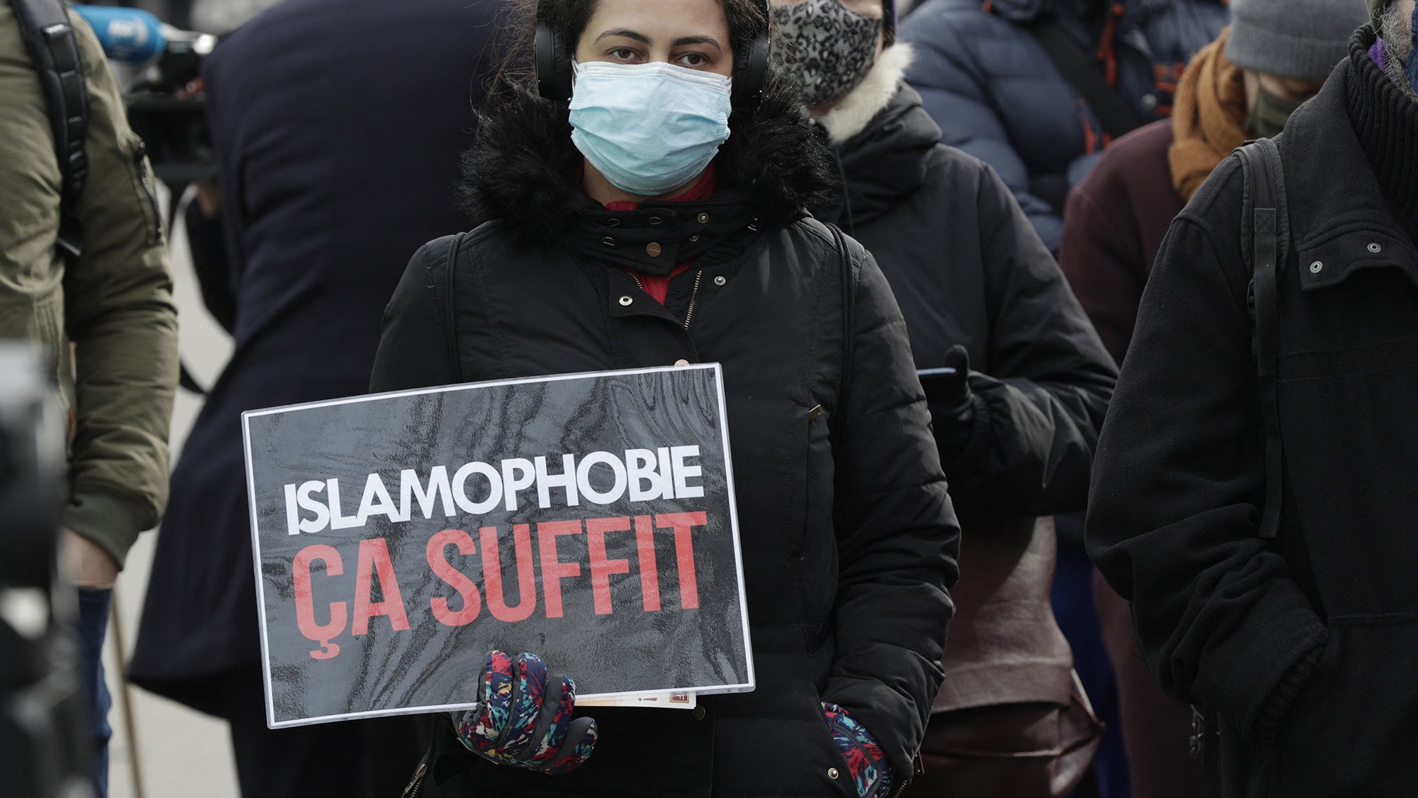 En la imagen se puede ver a una persona sosteniendo un cartel que dice "ISLAMOPHOBIE ÇA SUFFIT", lo que se traduce como "ISLAMOFOBIA BASTA YA". La persona lleva puesta una mascarilla y parece estar en un evento o manifestación, rodeada de otras personas que también usan mascarillas. El ambiente sugiere una expresión de protesta o de apoyo a una causa relacionada con la lucha contra la islamofobia.
