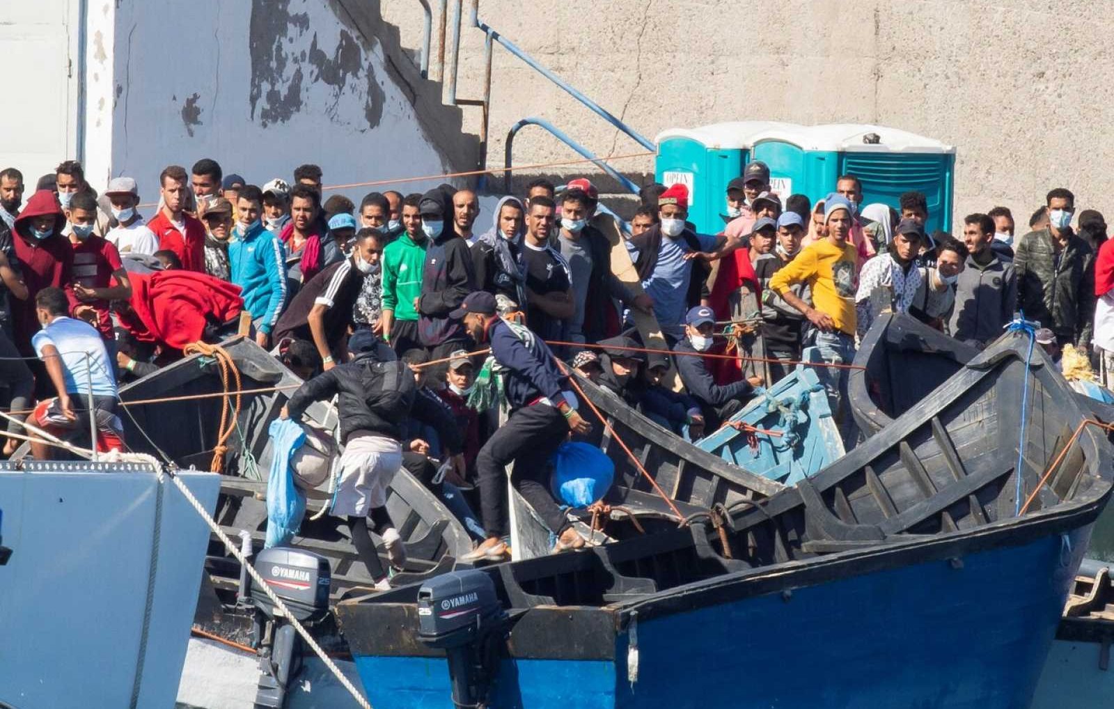 La imagen muestra un grupo de personas en un puerto o zona costera, donde hay varias embarcaciones. Muchas de las personas parecen estar en una o varias pequeñas embarcaciones de madera, mientras que otras están en la orilla o cerca de las embarcaciones. Se observa una multitud, con algunas personas vestidas en ropa casual, y parece que están en un momento de actividad o espera. Al fondo, hay estructuras de concreto y un baño portátil. La escena sugiere un ambiente de migración o búsqueda de oportunidades.