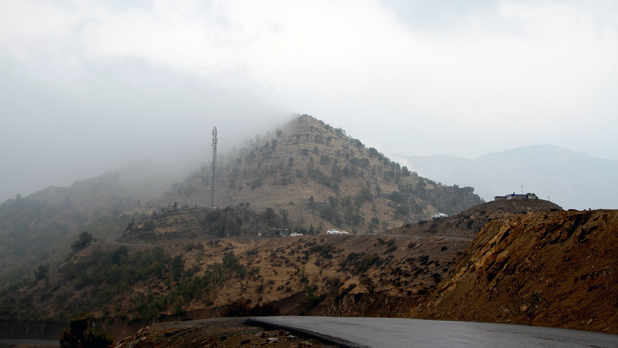La imagen muestra un paisaje montañoso con una carretera curvada en primer plano. Al fondo, se puede ver una colina cubierta de vegetación escasa y neblina, que le da un aspecto misterioso. En la parte superior de la colina, hay una torre de comunicación. El clima parece nublado y húmedo, creando un ambiente atmosférico y sombrío.