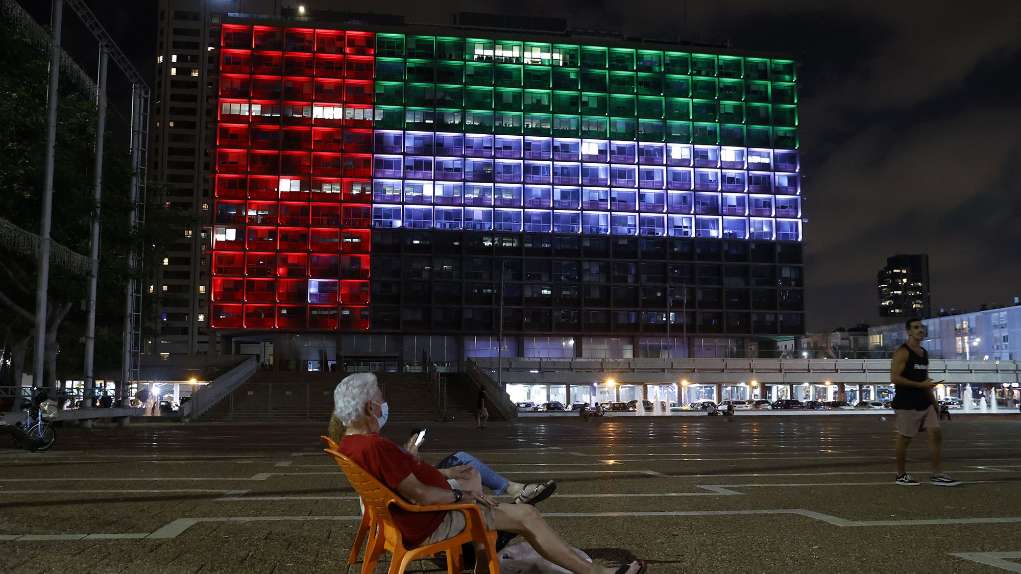En la imagen se puede ver a un hombre mayor sentado en una silla de plástico anaranjada, mirando su teléfono móvil. Detrás de él, se destaca un gran edificio iluminado que muestra los colores de la bandera de México: rojo, blanco y verde. Es de noche, y el ambiente parece tranquilo con algunas personas caminando en el área. La arquitectura del edificio se aprecia moderna, y las luces crean un contraste vibrante con el cielo nocturno.