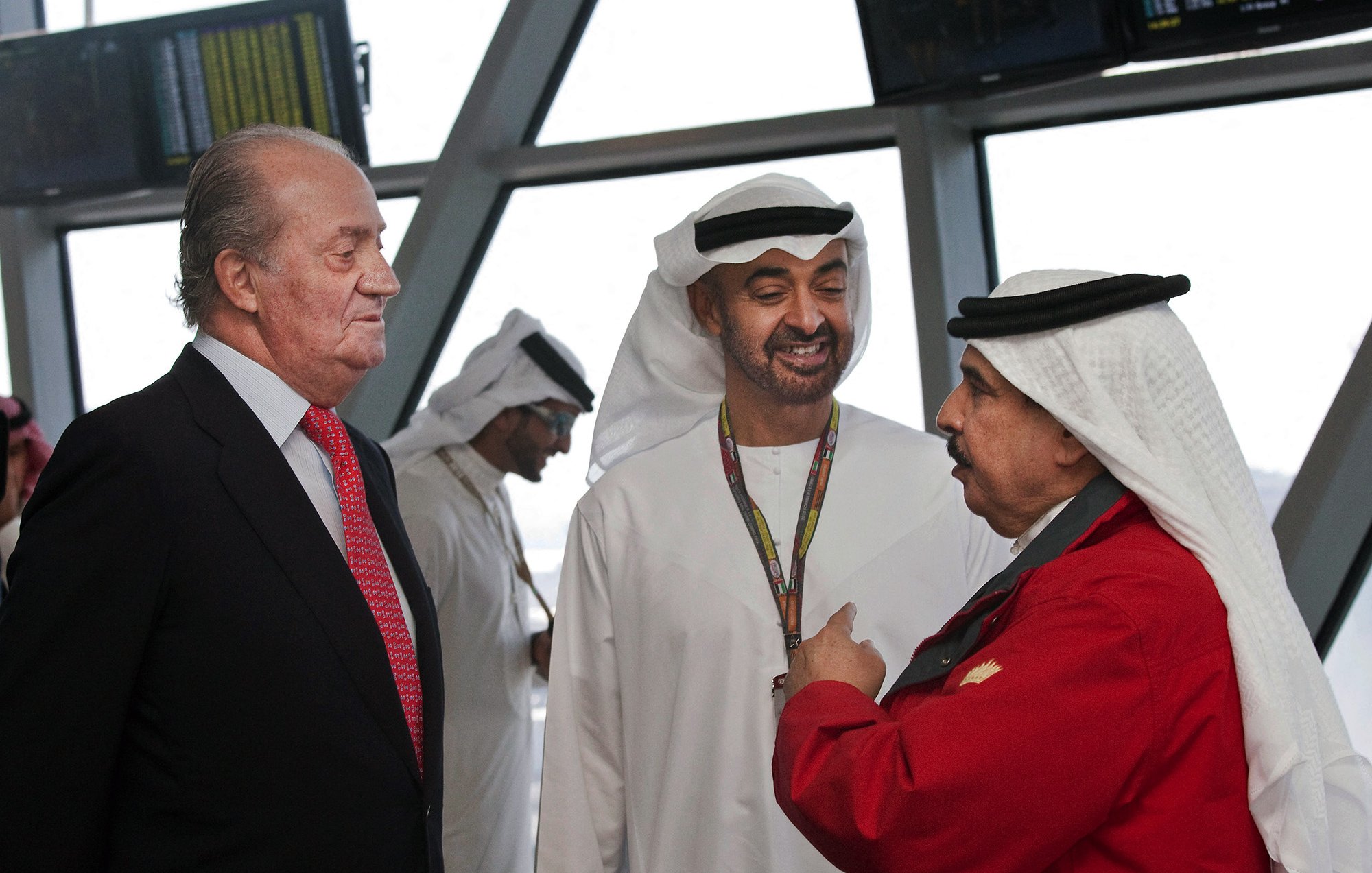 En la imagen se pueden ver a tres hombres conversando. Dos de ellos visten trajes tradicionales árabes, mientras que el tercero lleva un traje negro con una camisa blanca y una corbata roja. El entorno parece ser un aeropuerto o un lugar moderno. Los hombres están interactuando de manera amigable y hay pantallas en el fondo, lo que sugiere un ambiente ocupado y profesional.