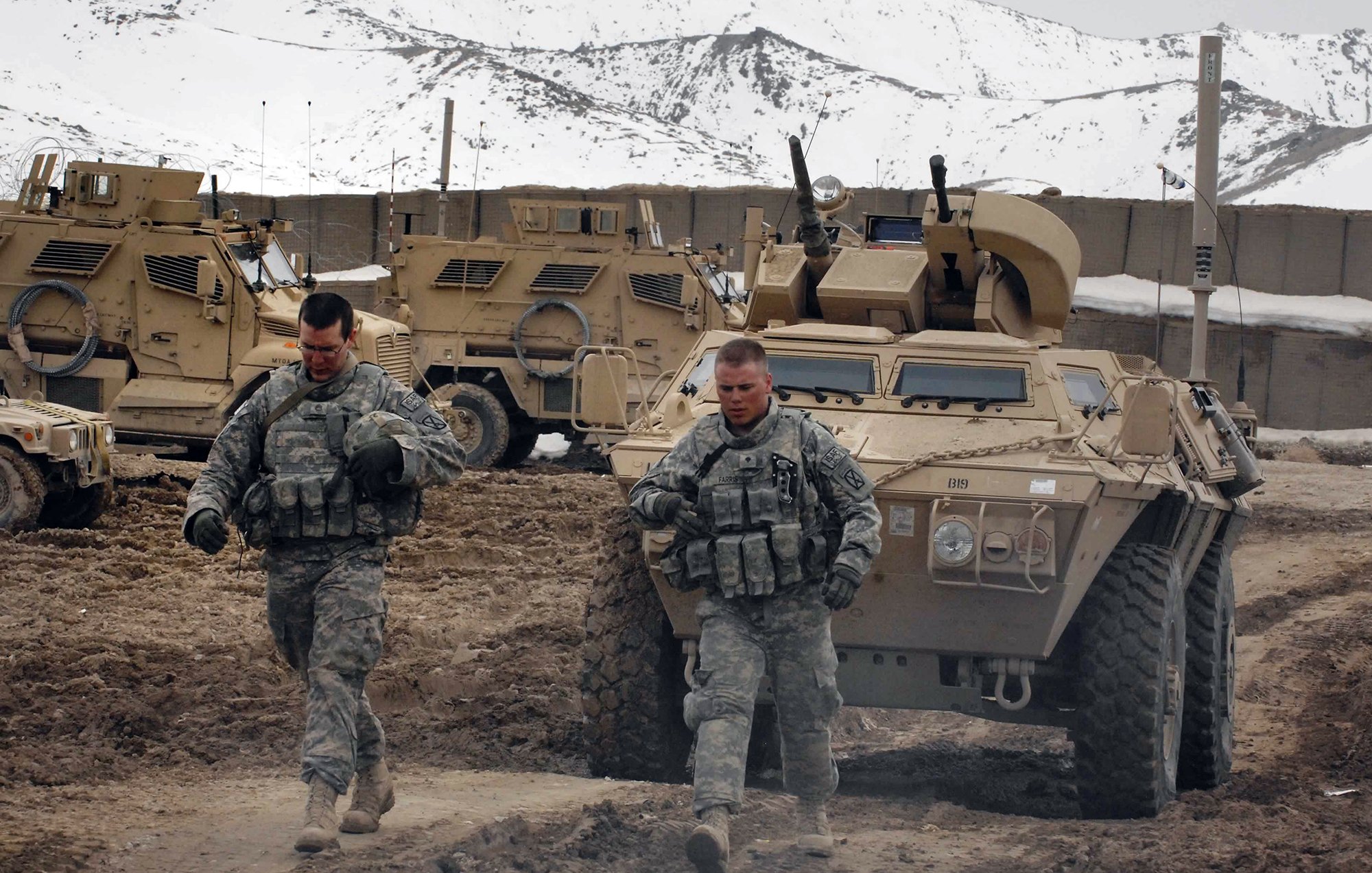 En la imagen se pueden ver a dos soldados caminando por un terreno irregular y arenoso. Están vestidos con uniformes militares y parecen estar en un entorno montañoso, con algunas unidades de vehículos blindados al fondo. La atmósfera es de actividad militar, con un paisaje que sugiere que están en una misión en un área remota. La nieve se puede observar en las montañas que rodean la escena.