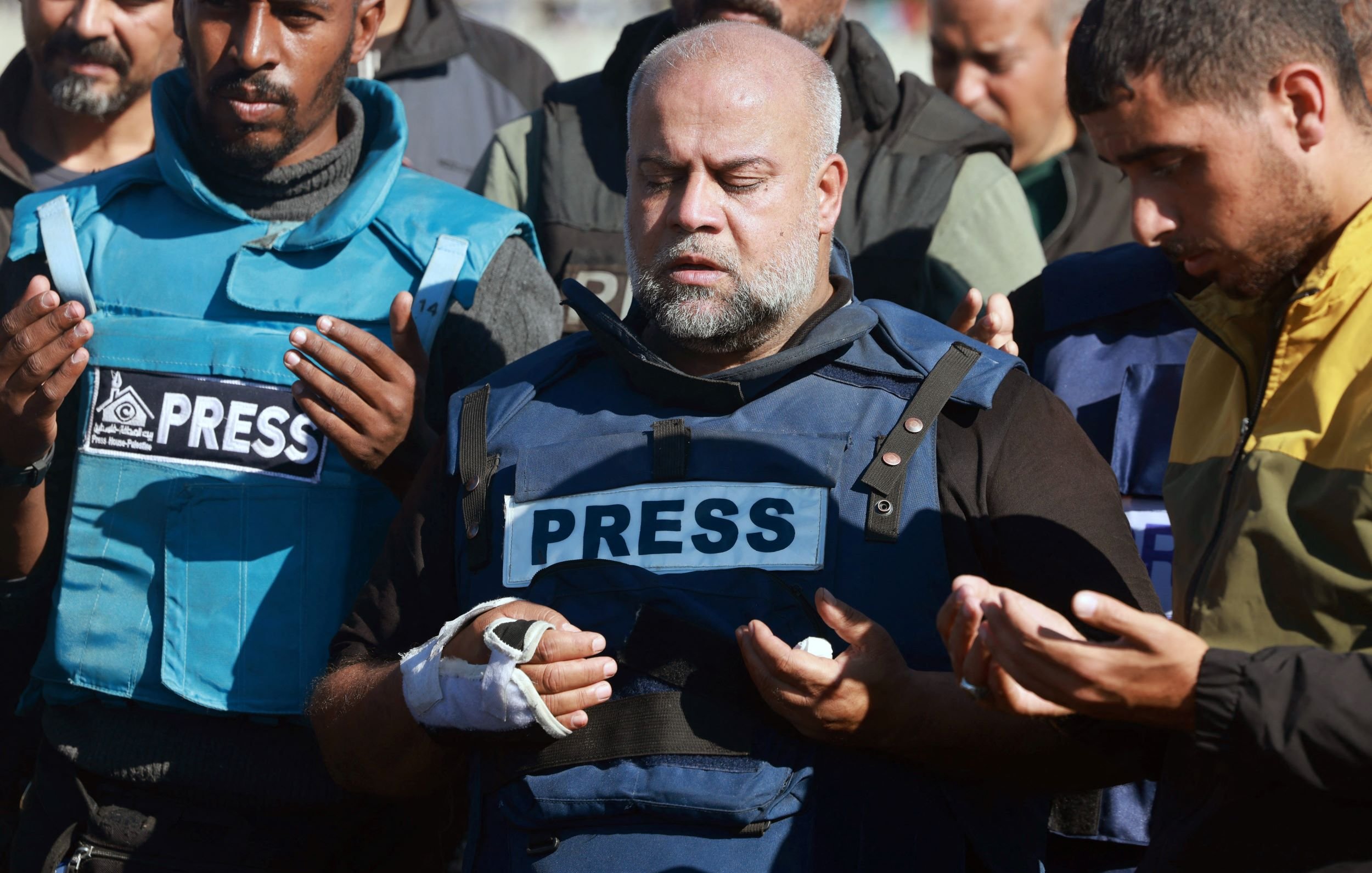 En la imagen, un grupo de personas está reunido, y se puede notar a un hombre en el centro que lleva un chaleco con la palabra "PRESS" (prensa) en grandes letras. Este hombre tiene una expresión de concentración y parece estar rezando. Su mano derecha está vendada. Los demás en el grupo también tienen una actitud similar, algunos con las manos levantadas en señal de oración. El ambiente sugiere un momento de solemnidad o de conmemoración.