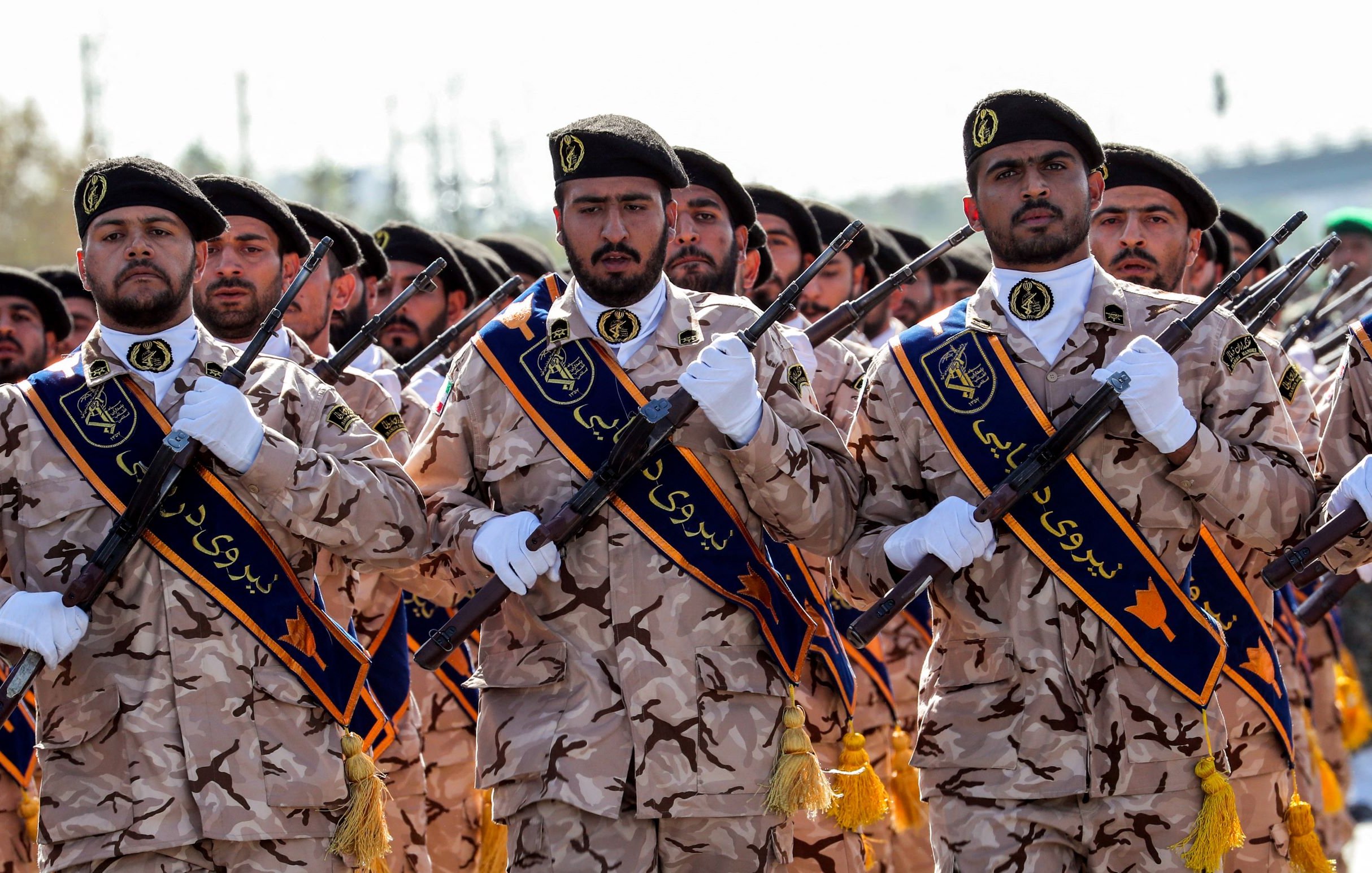 La imagen muestra a un grupo de soldados en un desfile militar. Están vestidos con uniformes de camuflaje y llevan distintivos en sus chaquetas. Los soldados sostienen rifles en posición vertical y parecen estar marchando con solemnidad. El ambiente es solemne y refleja un evento militar con un enfoque en la disciplina y la unidad.