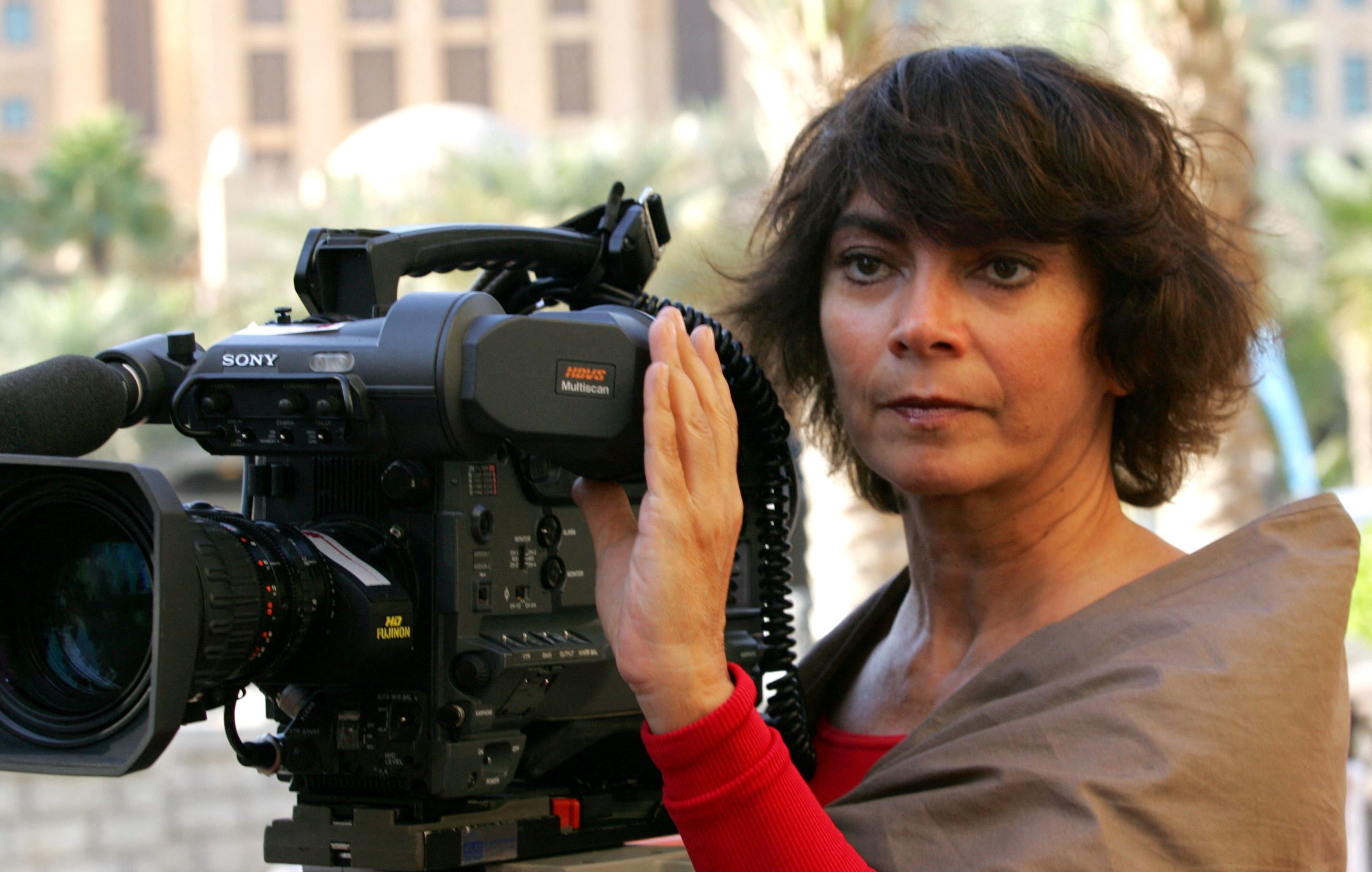 La imagen muestra a una mujer de cabello oscuro y rizado, que parece estar sosteniendo y operando una cámara de video profesional. Ella está mirando a la cámara con una expresión seria y concentrada. Viste una blusa color claro y tiene una capa o chal de un color más oscuro. Al fondo, se pueden ver edificios y un ambiente urbano. La escena sugiere que está en un evento o en el lugar de una grabación.