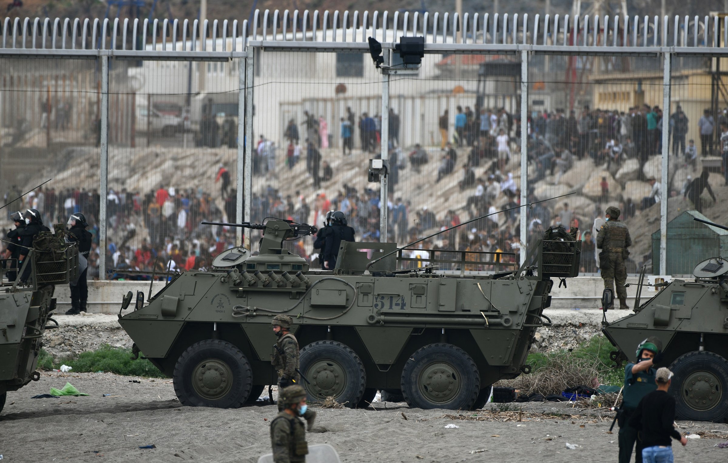 La imagen muestra un contexto de tensión, probablemente en una zona de conflicto o crisis. Se pueden ver vehículos militares blindados en la parte frontal, con soldados a bordo y personal militar en el entorno. Al fondo, hay una multitud de personas que parecen estar frente a una valla alta, indicando una separación entre diferentes grupos. El ambiente es de intensa actividad, posiblemente con indicios de enfrentamientos o una situación de disturbio. La escena evoca un fuerte sentido de vigilancia y seguridad militar.