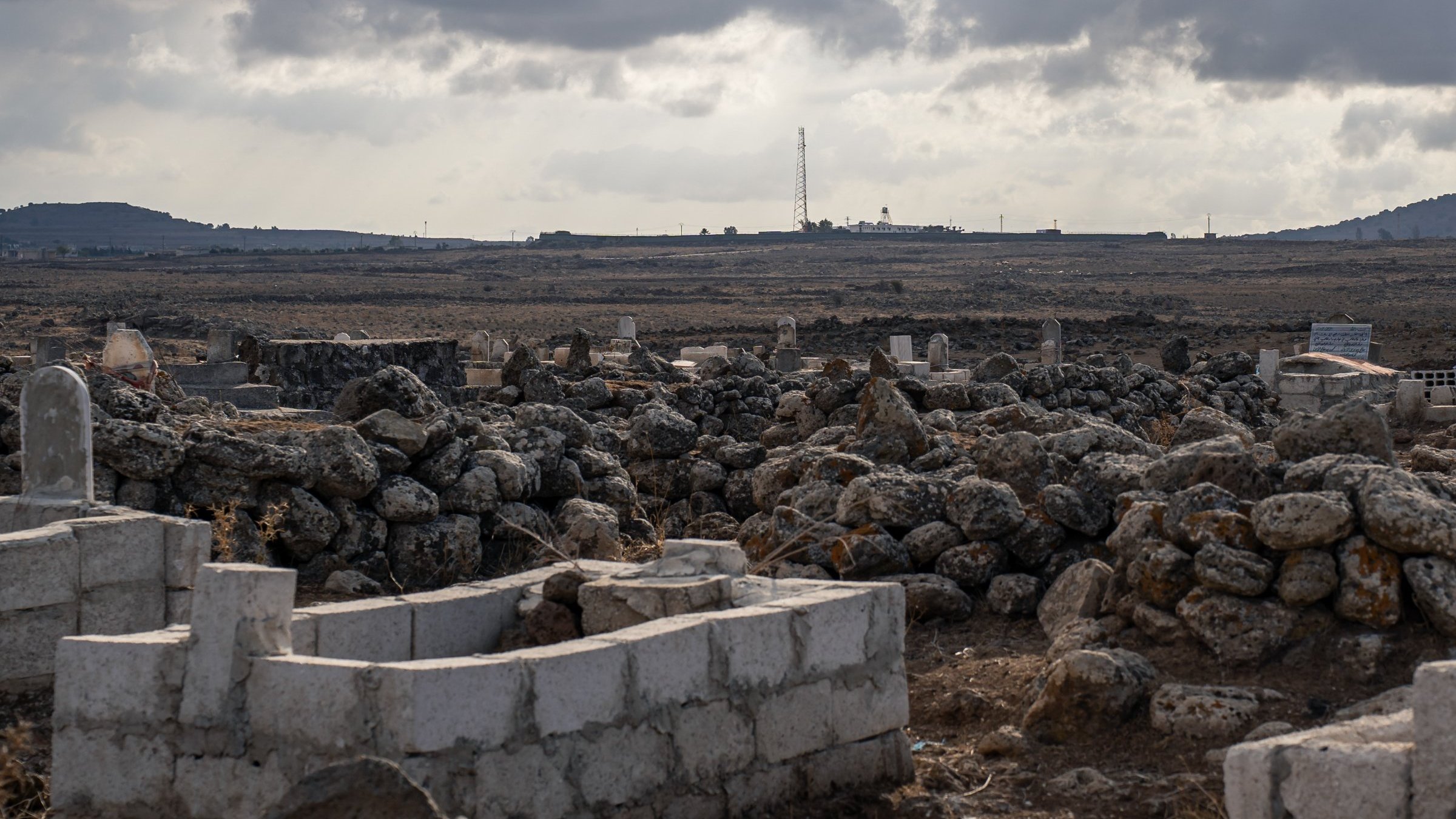 Un paisaje árido con tumbas y rocas, bajo un cielo nublado y distante, se ve una torre.