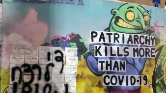 La imagen muestra un mural de arte urbano. En el lado izquierdo, hay una figura caricaturesca de un ser verde con una gran sonrisa, que sostiene un cartel que dice "PATRIARCHY KILLS MORE THAN COVID-19". El fondo tiene un paisaje colorido con nubes y algunos elementos abstractos. A la izquierda del mural, hay graffiti que parece ser un mensaje adicional, aunque no es claramente legible. La combinación de colores y estilos resalta un enfoque crítico hacia el patriarcado, comparándolo con los efectos de la pandemia.
