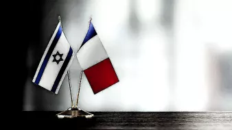 La imagen muestra dos banderas pequeñas en un soporte: una de Israel, que presenta la estrella de David en un fondo blanco y azul, y otra de Francia, con los colores azul, blanco y rojo dispuestos verticalmente. Las banderas están colocadas sobre una superficie oscura, y el fondo es difuso y claro, lo que resalta las banderas.
