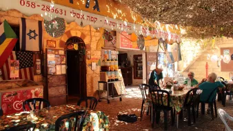 La imagen muestra un ambiente acogedor y colorido de un café o restaurante al aire libre. Hay mesas y sillas negras distribuidas sobre un suelo pavimentado, y la decoración incluye manteles con estampados florales. En el fondo, se pueden ver varias banderas, incluyendo la de Israel, colgadas en las paredes de piedra. El espacio está cubierto por una especie de toldo que filtra la luz, creando un ambiente agradable. Algunas personas están sentadas en las mesas, disfrutando del lugar.