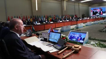 En la imagen se puede ver a un grupo de personas reunidas en una sala de conferencias. Al frente, hay un hombre sentado en una mesa, supervisando la reunión. Detrás de él, se observa una fila de delegados de diferentes países, muchos de los cuales están sentados al lado de mesas decoradas con banderas nacionales. También hay pantallas detrás de ellos mostrando imágenes o videoconferencias de otros participantes. La sala está adornada con arreglos florales y hay un ambiente formal y profesional, característico de una cumbre o conferencia internacional.